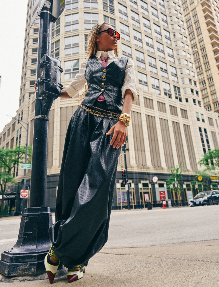 A woman stands on a city street wearing a long black wide-leg skirt with a gold waist detail, a black vest over a cream shirt, and gold accessories. Tall city buildings are visible in the background.