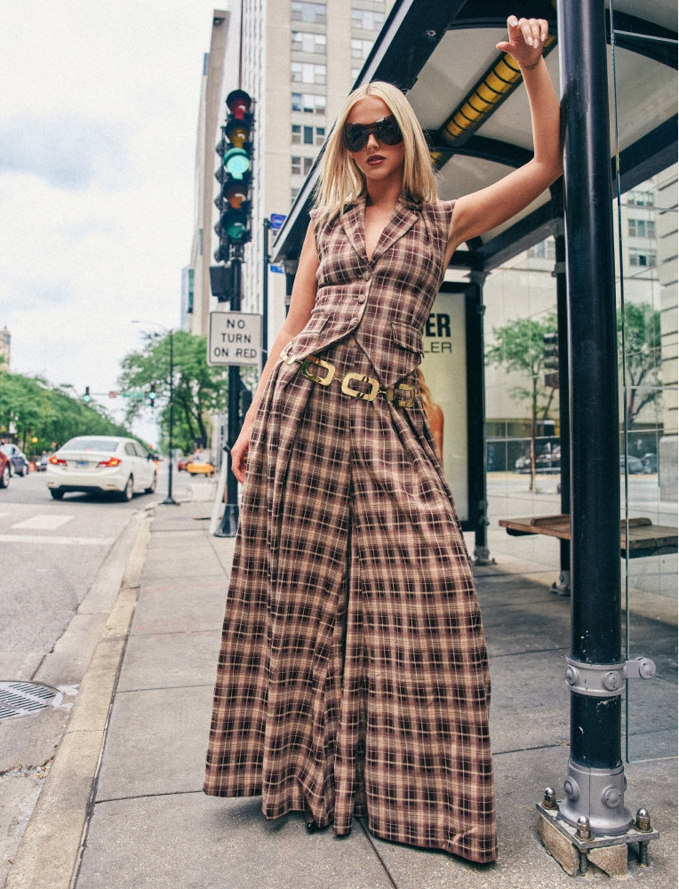A woman wearing a brown and beige plaid sleeveless jumpsuit with a matching belt stands on a city sidewalk next to a bus stop shelter.