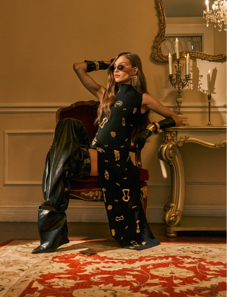 A woman poses in an ornate vintage room wearing a black ribbed sleeveless dress with gold key motifs, black leather boots, and a black leather jacket draped over a chair.