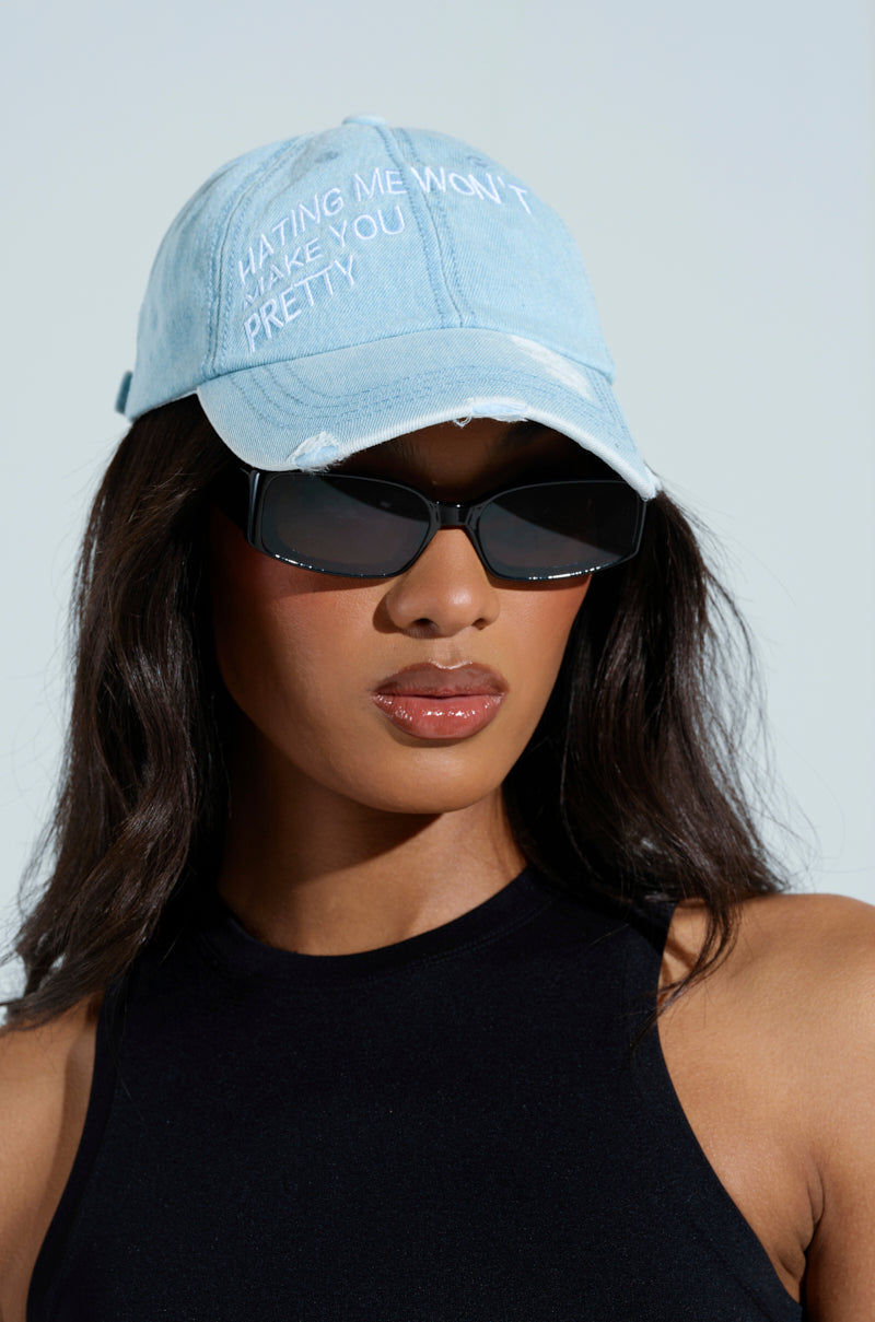 Wearing a light blue STRAIGHT FACTS SNAPBACK embroidered with "HATING ME WON'T MAKE YOU PRETTY," dark sunglasses, and a sleeveless black top, a person poses against a light background.