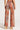 Someone is dressed in the TOO HOT TO CARE PANT from the AKIRA Label, featuring high-waisted, wide-leg pants with a shimmery metallic base and a vibrant pattern in shades of orange, brown, and purple. They are also wearing high-heeled sandals. The photo captures the back view of this stunning outfit.