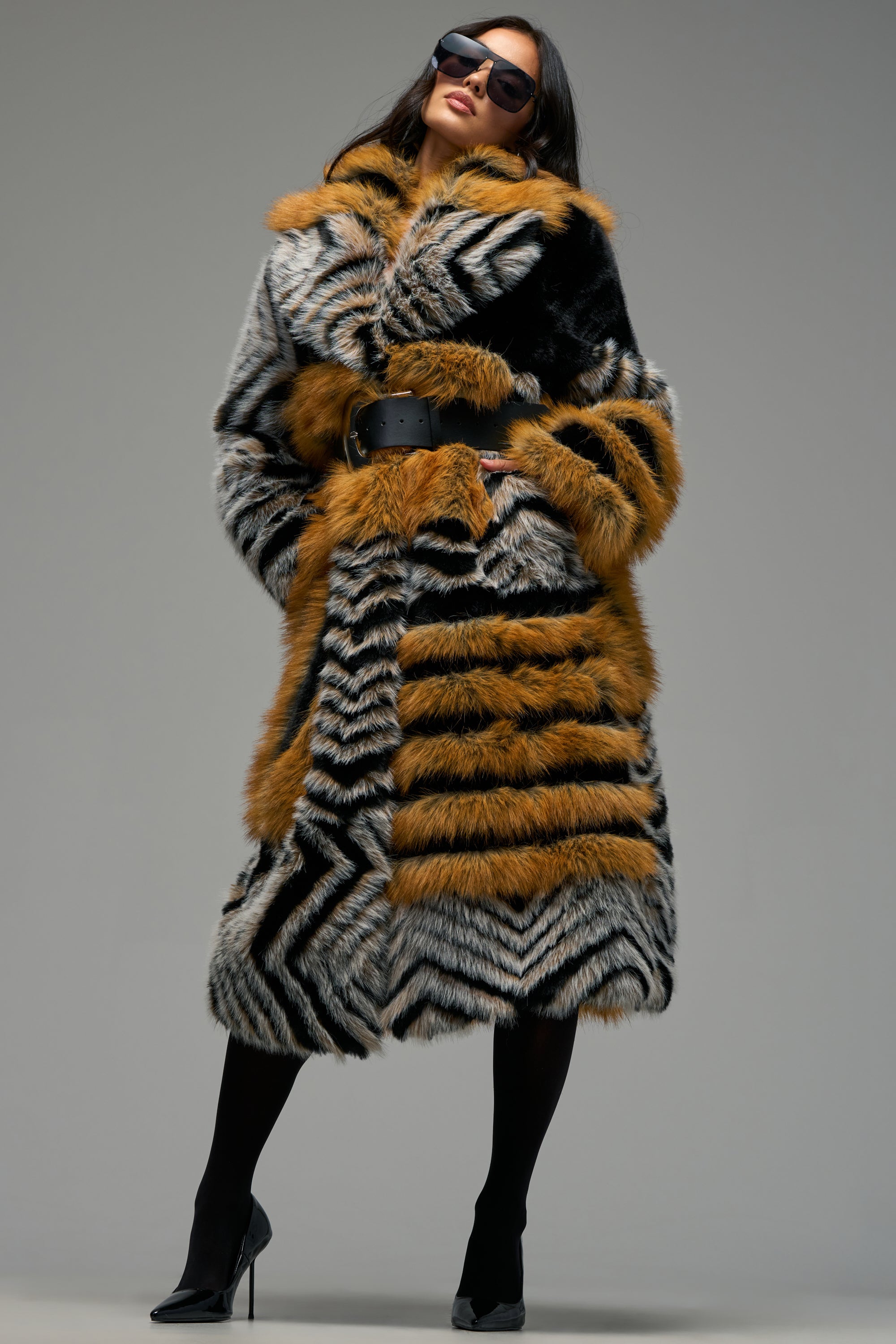 Wearing the PHANTOM PATCHWORK FAUX FUR, a woman stands confidently against a neutral background. This statement winter coat features bold black, white, and orange striped patterns in a longline silhouette with a belted waist for added definition. Paired with sunglasses, black tights, and high-heeled shoes, the striking color combination ensures all eyes are drawn to her unforgettable look.