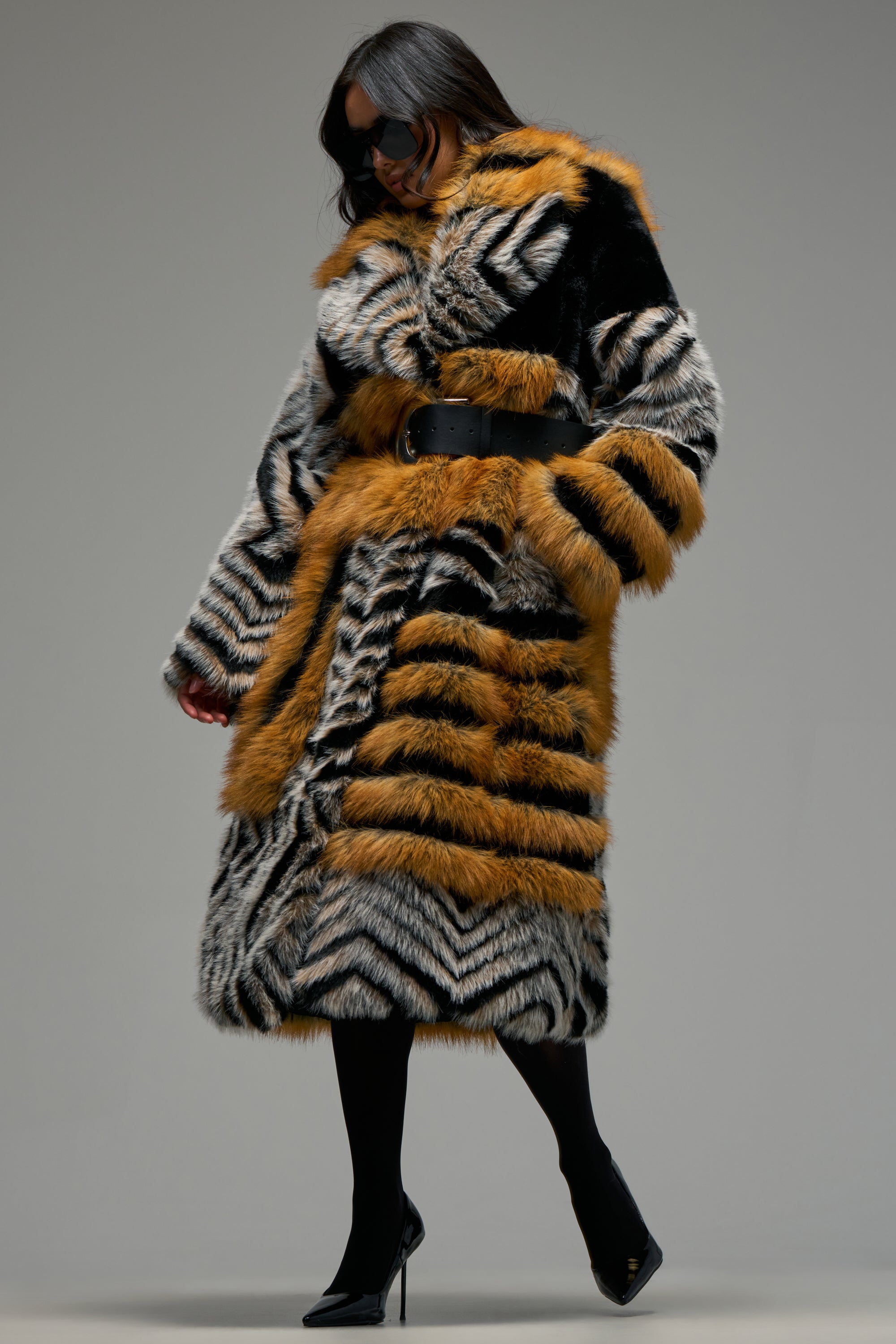 A person wearing sunglasses and the PHANTOM PATCHWORK FAUX FUR—a longline faux fur coat featuring bold black, white, and orange stripes, belted at the waist—stands against a plain background. This striking statement winter coat is paired with black tights and high heels, creating a bold, fashion-forward look.