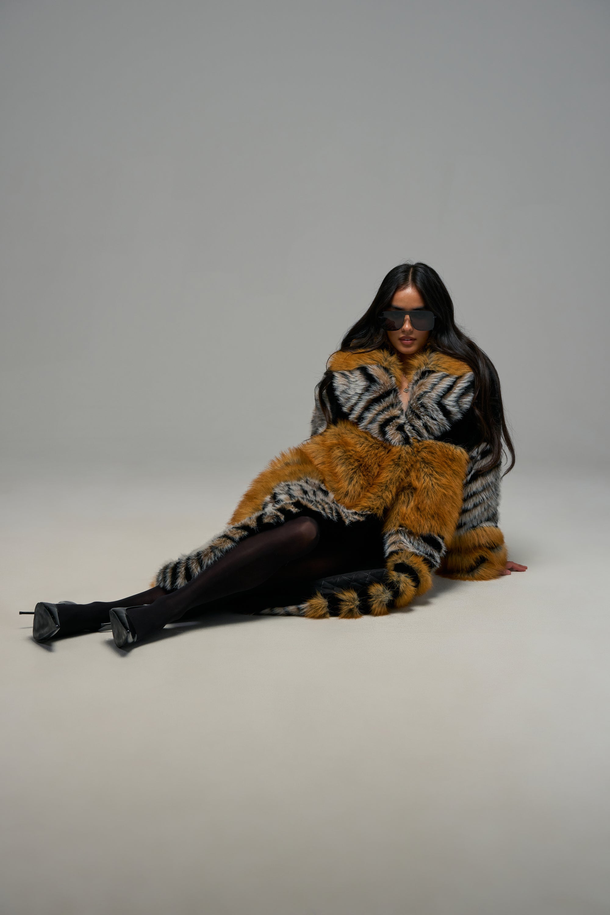 A person with long dark hair wearing sunglasses, black tights, and heels sits on the floor against a plain light background, showcasing the PHANTOM PATCHWORK FAUX FUR statement winter coat in striped orange and gray.