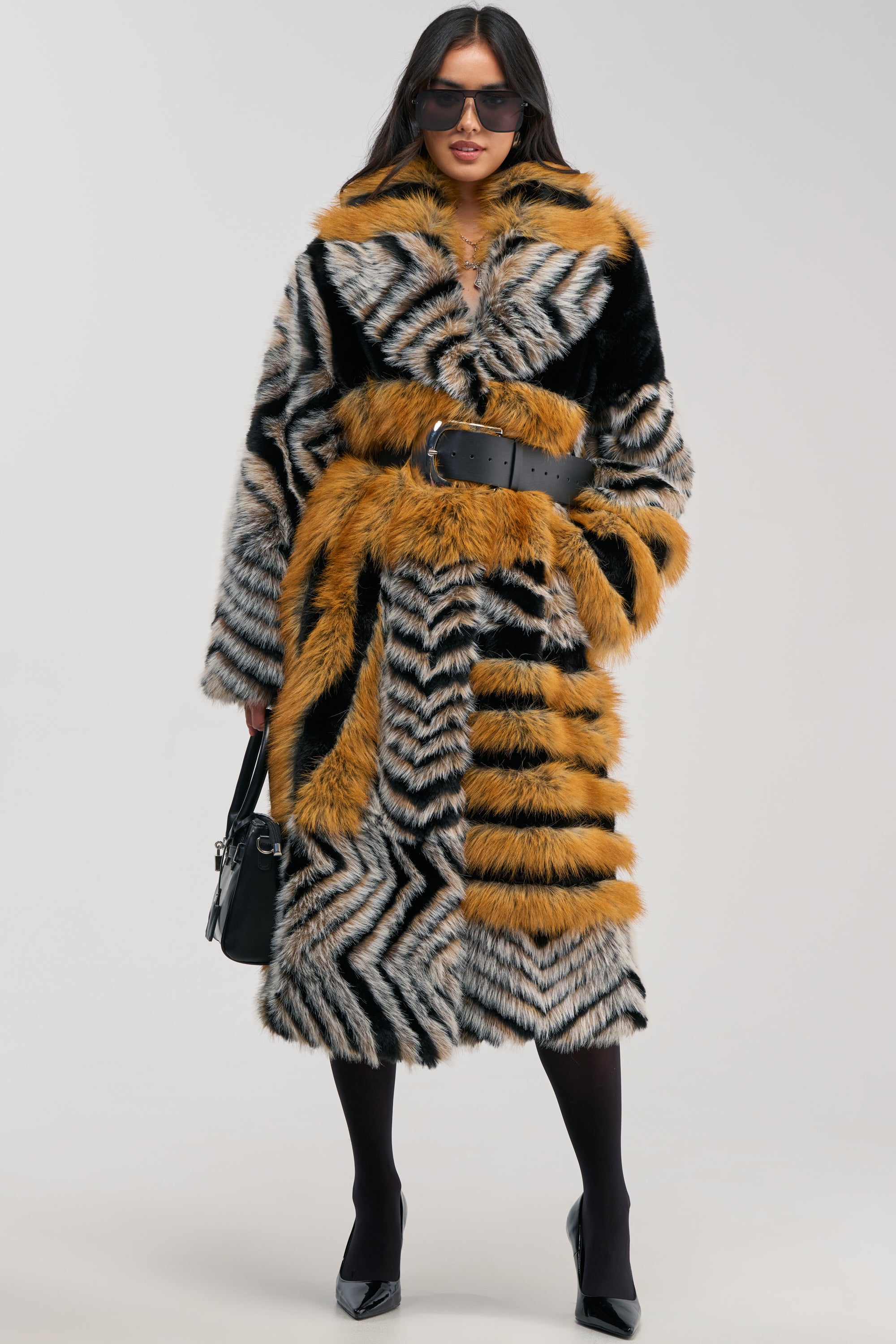 A woman models the PHANTOM PATCHWORK FAUX FUR winter coat, showcasing its bold black, gray, and orange stripes and a cinched black belt. She completes the ensemble with black tights, pointed heels, sunglasses, and a chic black handbag while posing against a plain background.