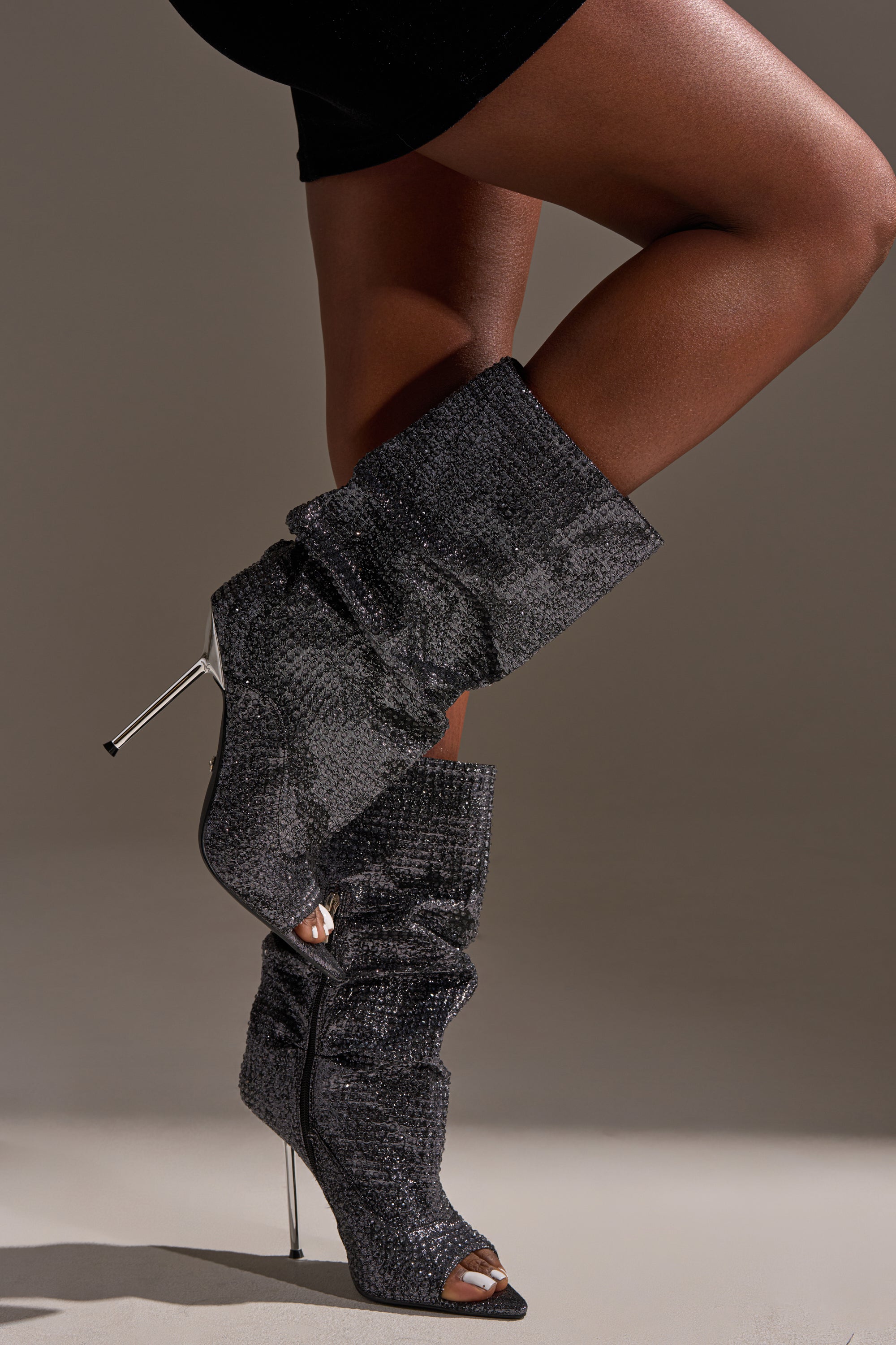 A person in a black mini skirt stands with one leg crossed over the other, wearing AZALEA WANG PHOTINE BLACK MULTI SPARKLE OPEN TOE STILETTO BOOTIES against a neutral background.