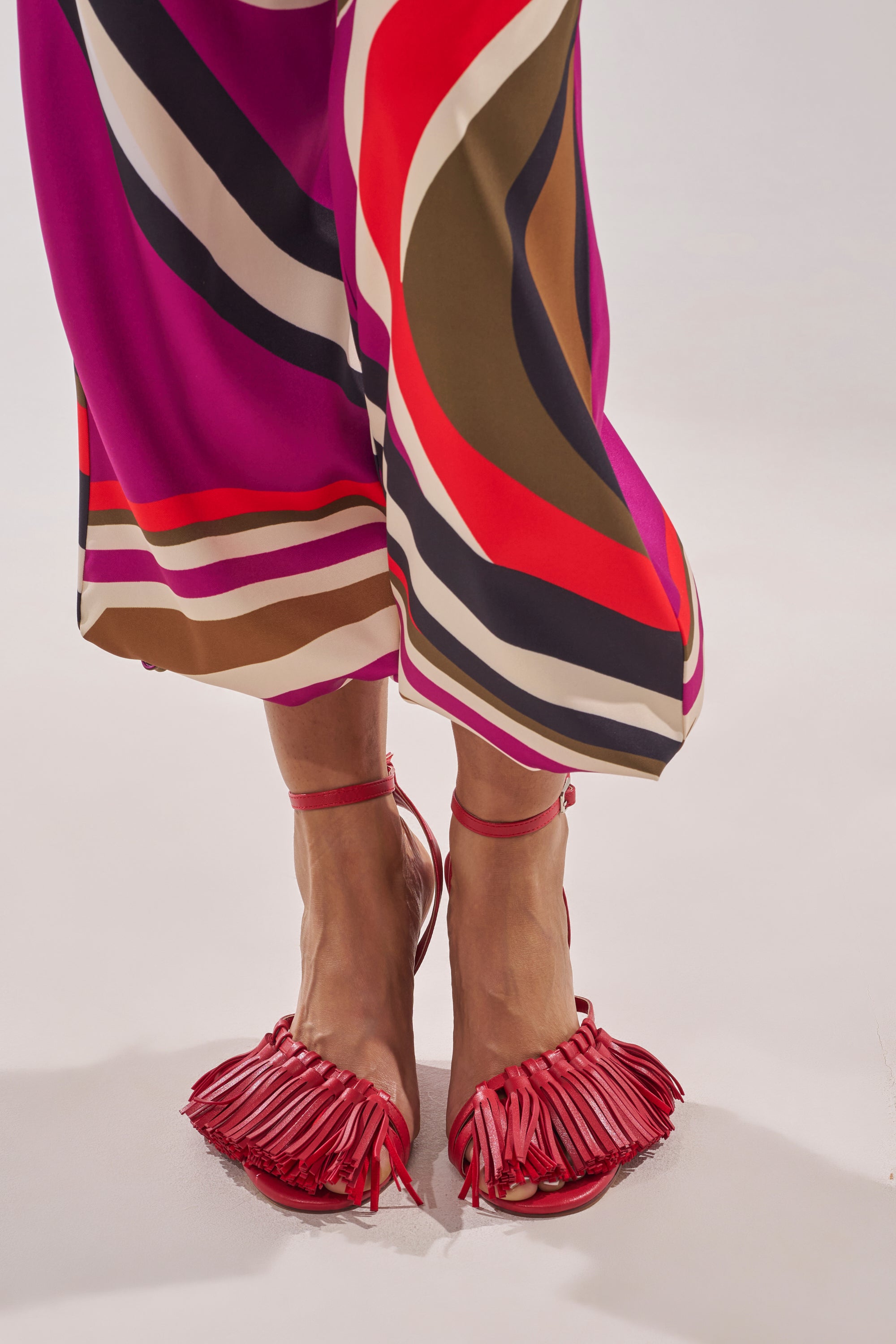 Wearing geometric-print pants in shades of pink, red, brown, and black, a person pairs them with vibrant AZALEA WANG PHRIXOS RED WEDGE FRINGE PU SANDALS, standing against a light background. 1