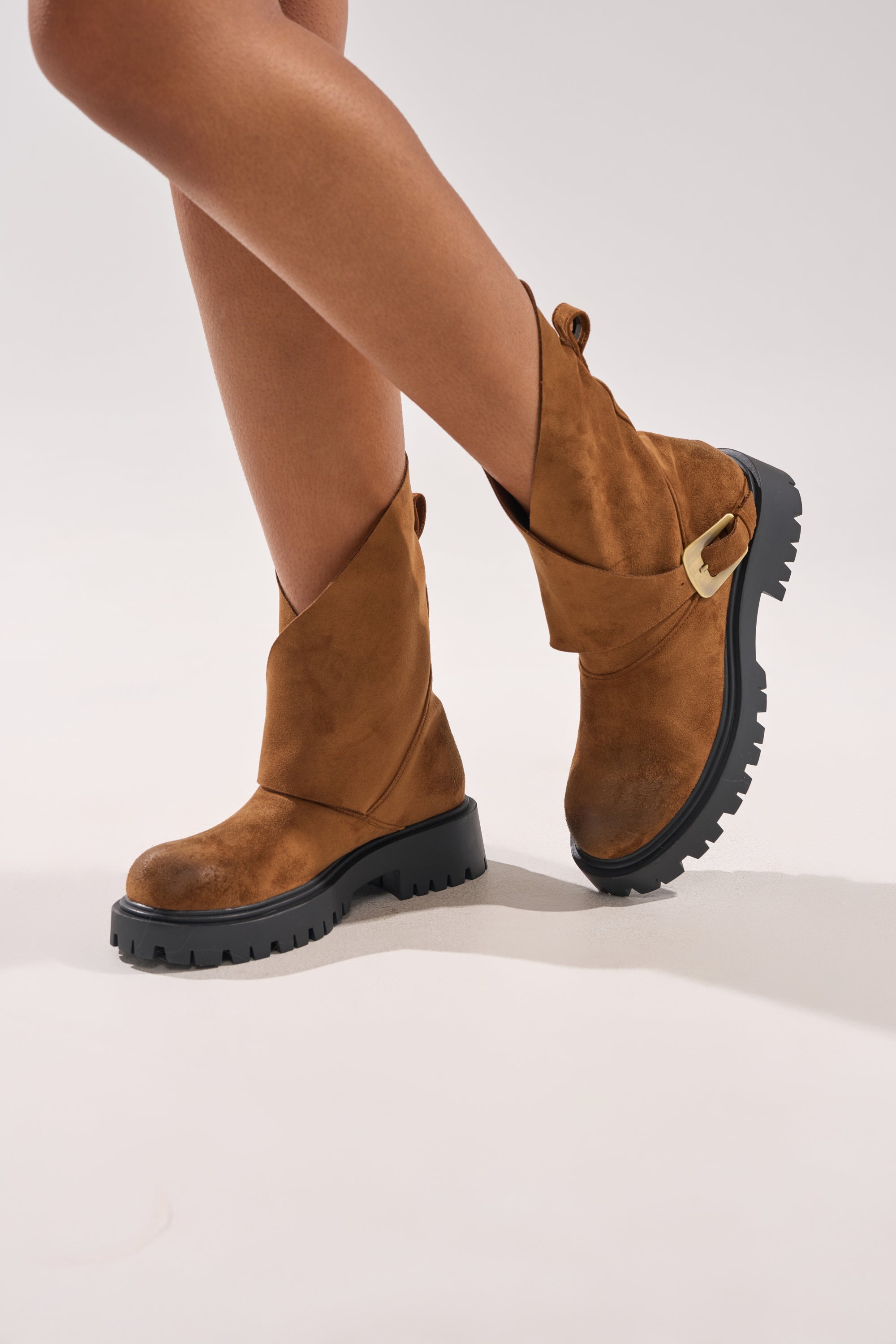 A person models the AZALEA WANG PHRIXUS Brown Chunky Short Shaft Faux Suede Boot, featuring a mid-calf height, gold buckle detail, and black platform lug sole, standing with one leg crossed on a light background.