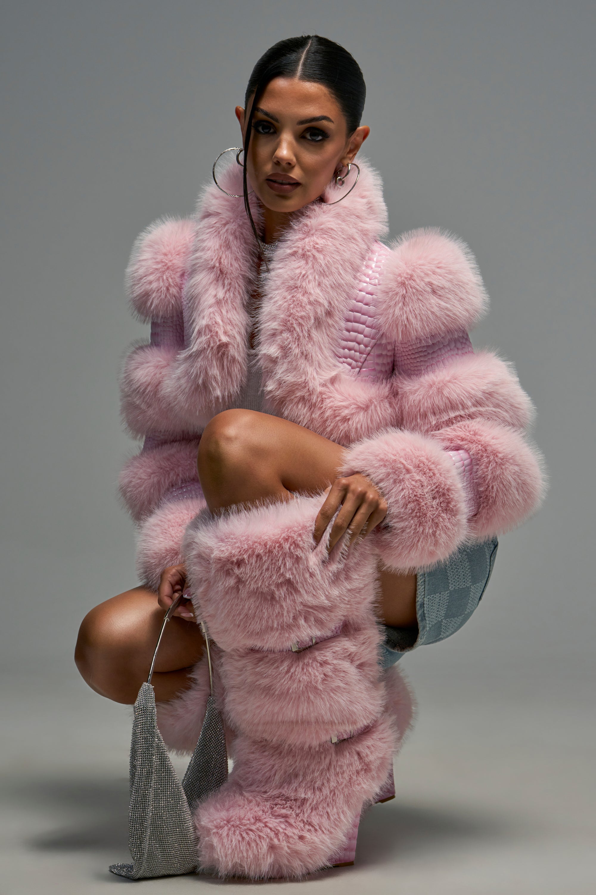 A woman in a pink faux fur jacket and matching AZALEA WANG PILLOW TALK PINK WEDGE BOOT knee-high boots kneels while holding a small, metallic silver handbag.