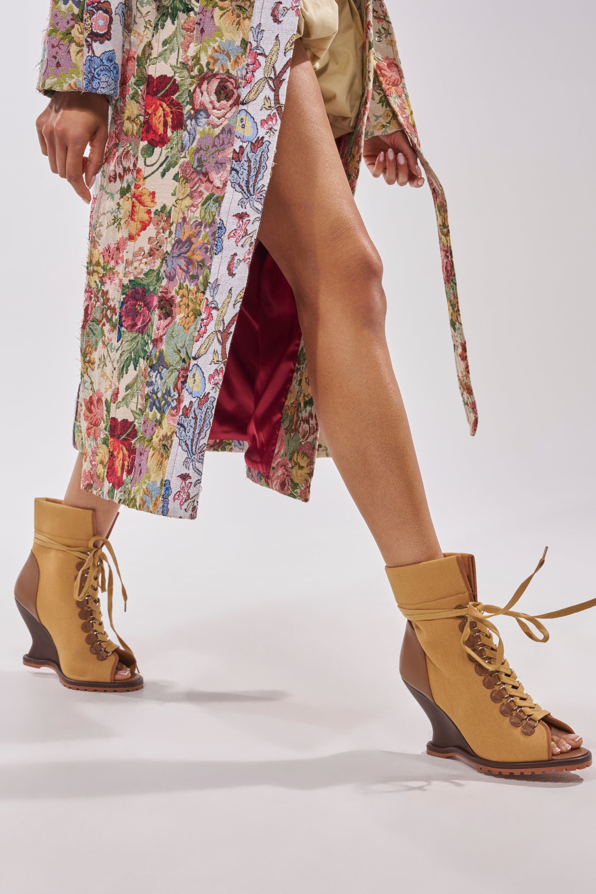 A person in statement streetwear walks forward, sporting AZALEA WANG PINGREE BROWN OPEN TOE WEDGE BOOTIES and a vibrant floral coat with red satin lining. The image highlights their legs and shoes as one foot is slightly lifted. 0