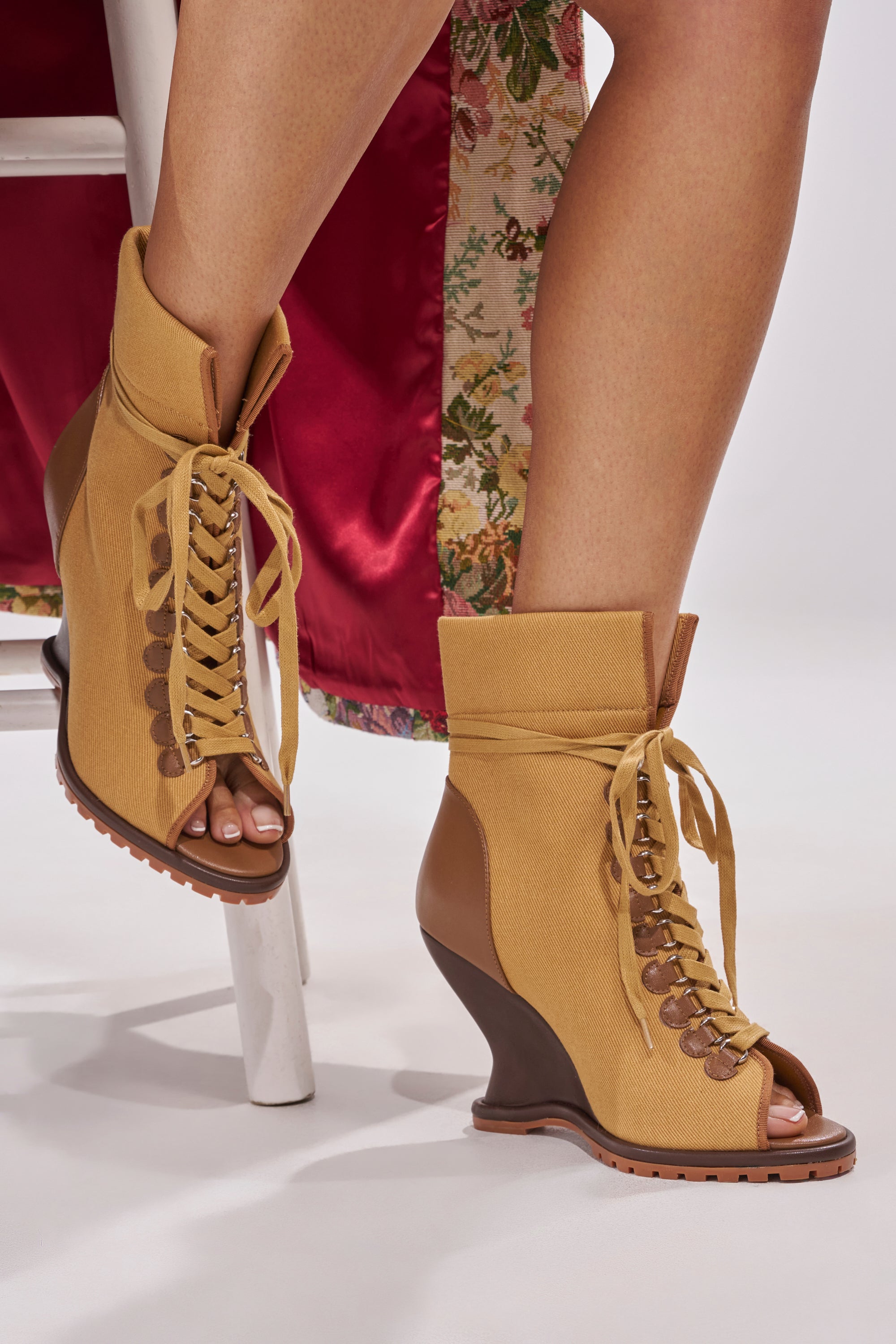 A close-up of a person wearing the AZALEA WANG PINGREE BROWN OPEN TOE WEDGE BOOTIE beside a chair draped with floral and red fabric—ideal streetwear booties for any casual outfit. 2