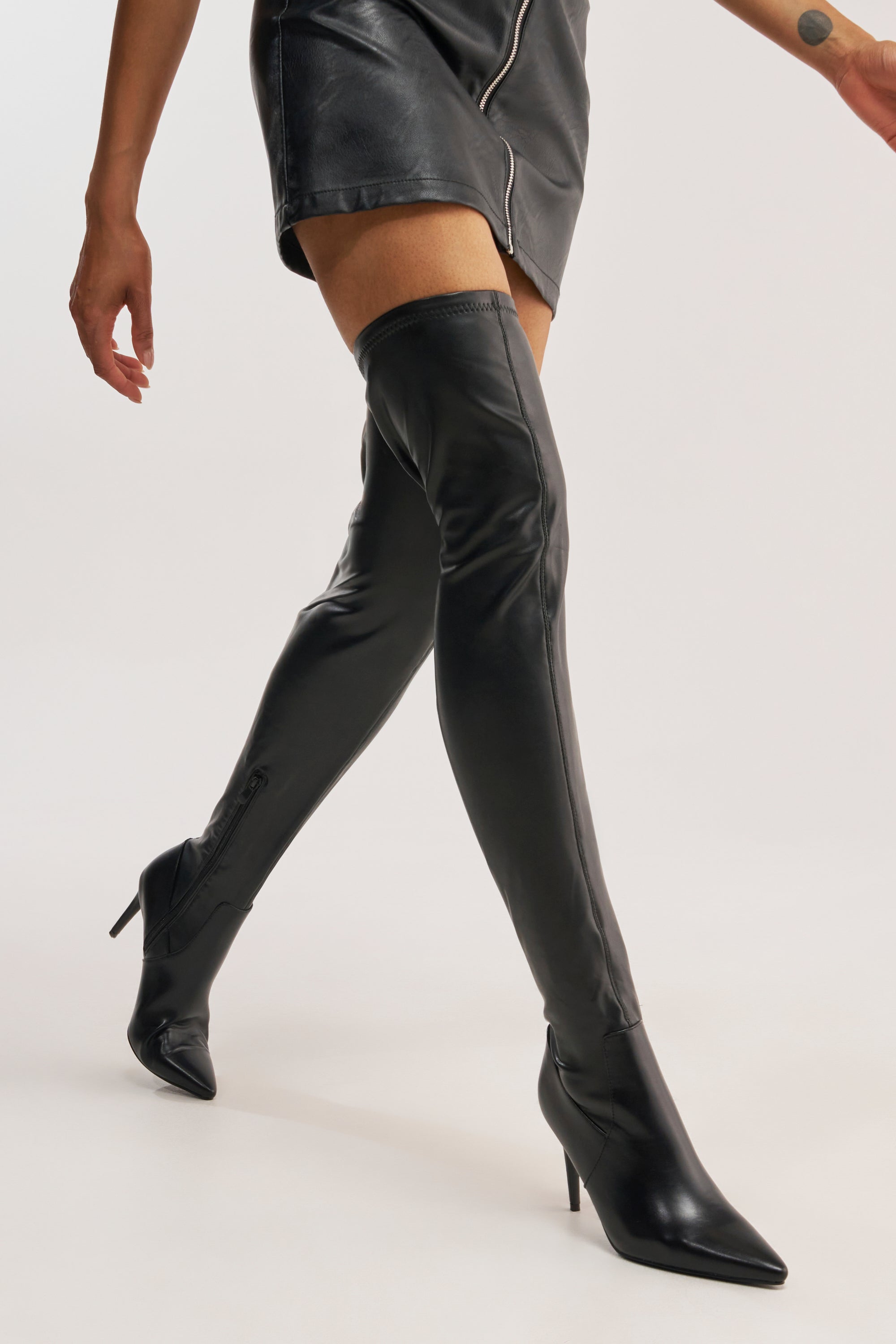 A person walks with only their lower body visible, wearing a black leather mini skirt and AZALEA WANG PINUP BLACK STILETTO BOOT against a plain background.