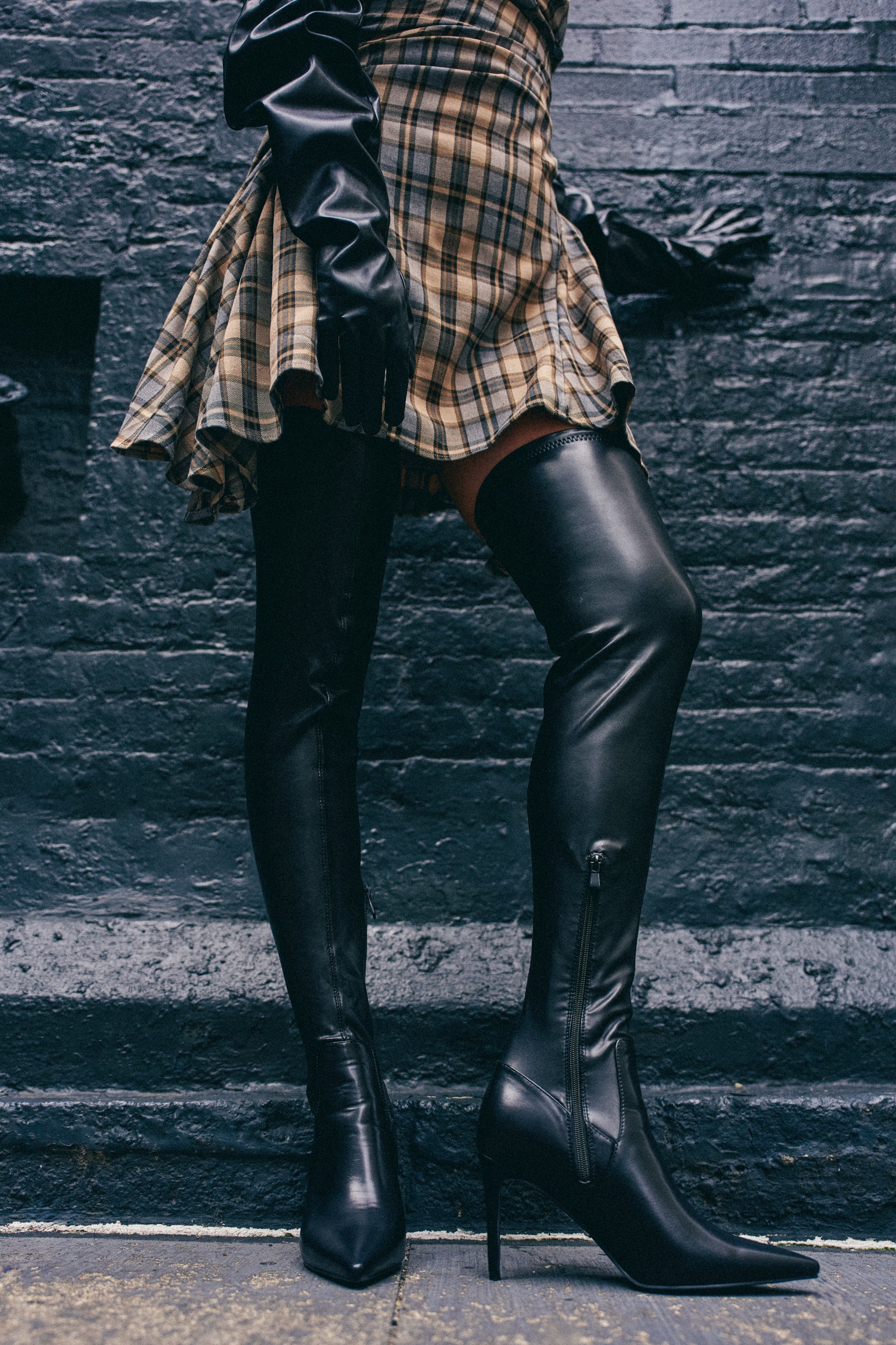 Wearing a plaid skirt, black leather gloves, and the AZALEA WANG PINUP BLACK STILETTO BOOT, a person stands against a dark brick wall with only the lower half of their body visible.