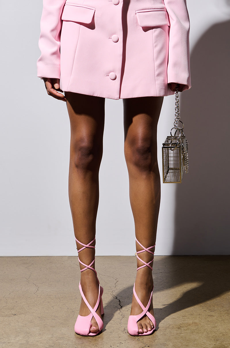A person stands against a plain backdrop, showcasing the lower half of their outfit, which features an oversized light pink blazer paired with strappy high heels in Pirate Pink from the PIRATE PINK PUMP collection. They are accessorizing with a small, transparent box-like handbag that has a metal chain.