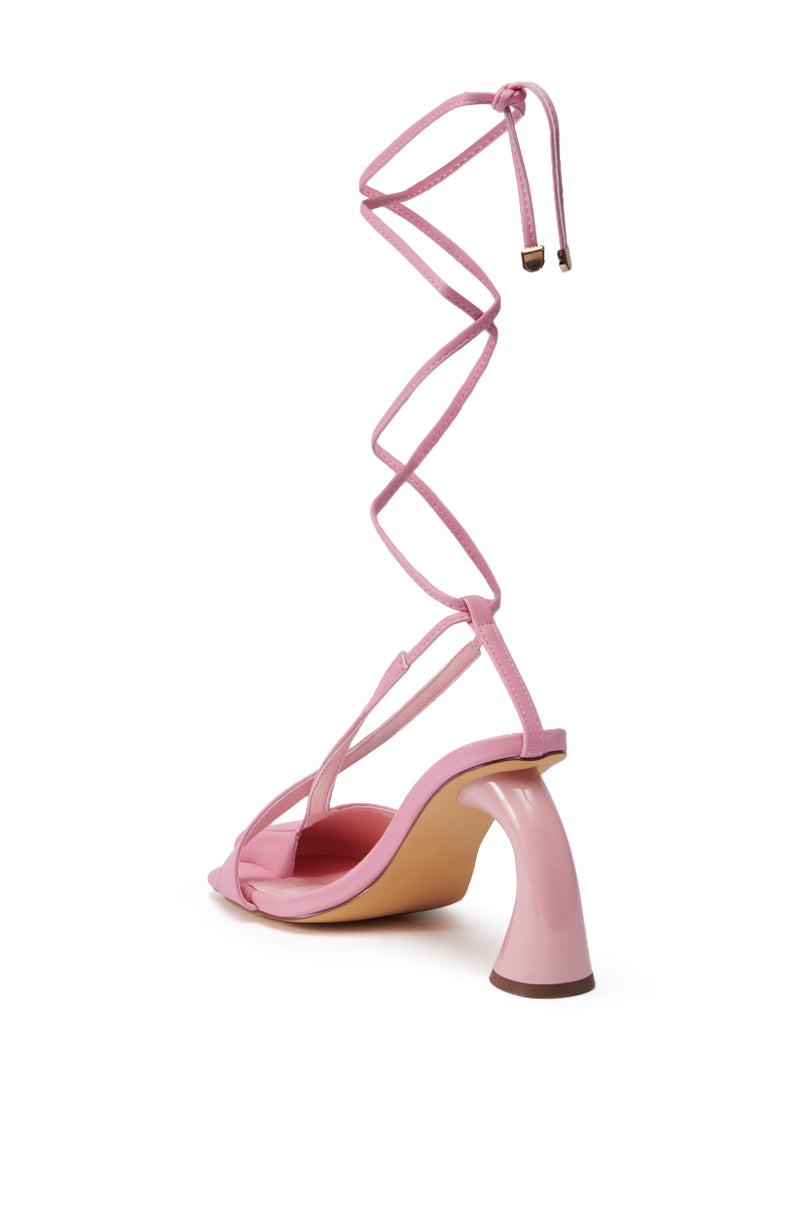Introducing the PIRATE PINK PUMP: a fashionable high heel sandal boasting a distinctive curved chunky sculpted heel. This shoe captures the essence of the balletcore trend with its crossed straps and extended wraparound ankle ties, achieving an elegant and modern aesthetic. The vibrant pink color is perfectly complemented by a light tan sole.