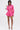 A woman poses against a plain backdrop in the LOOK THIS WAY SHINY SHORT, a metallic pink long-sleeve romper with flared shorts, styled with a pearl-studded round handbag and silver high-heeled sandals.