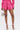 A woman wears the LOOK THIS WAY SHINY SHORT in hot pink with a matching top, pairs it with silver high-heeled sandals and a round silver handbag. She poses against a plain white background.
