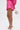 A person in profile wears the LOOK THIS WAY SHINY SHORT, a shiny hot pink high-rise skirt with a matching blouse, clear rhinestone-embellished heels, and holds a round silver beaded purse against a plain white background.