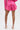 A person stands with their back to the camera, showing off the LOOK THIS WAY SHINY SHORT in hot pink metallic fabric, paired with a matching top and strappy high-heeled sandals against a plain white background.