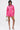 A woman poses in the LOOK THIS WAY SHINY SHORT—a vibrant pink, long-sleeve romper with a deep V-neck and pleated high-rise shorts—paired with strappy sandals and a small round handbag.