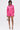A woman poses against a white background in the LOOK THIS WAY SHINY SHORT, a bright pink, long-sleeved romper with a pleated skirt and keyhole neckline. She completes the look with a beaded handbag and silver high-heeled sandals.