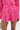 A person in the LOOK THIS WAY SHINY SHORT—shiny, bright pink high-waisted shorts with pleats and two buttons—paired with a matching pink top and a small silver bag for a bold, coordinated look.