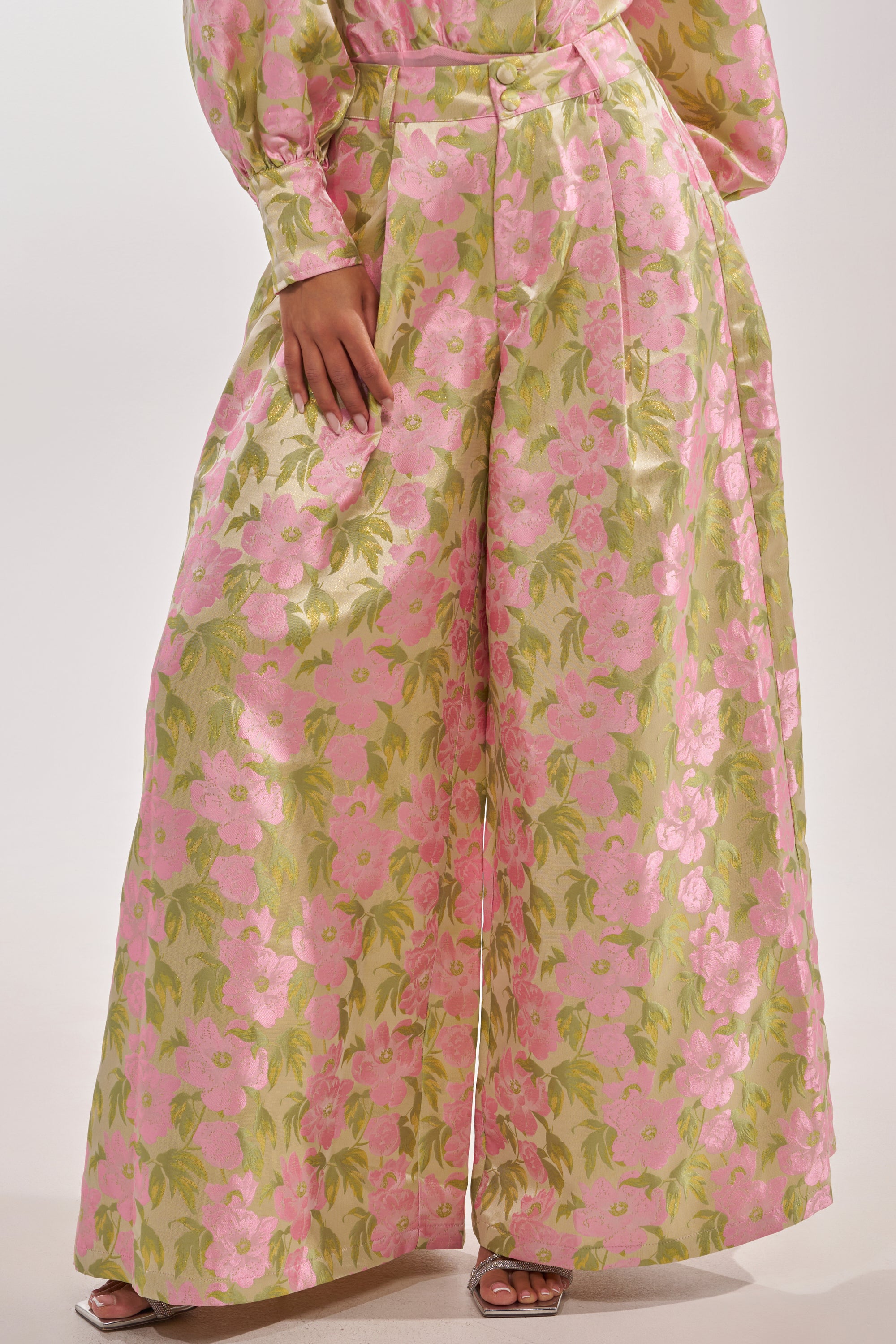A person wearing the OVER IT FLORAL TROUSER, featuring a wide-leg fit, high-rise waist, and floral print, with a matching top and silver open-toe heels. The background is plain white. 1