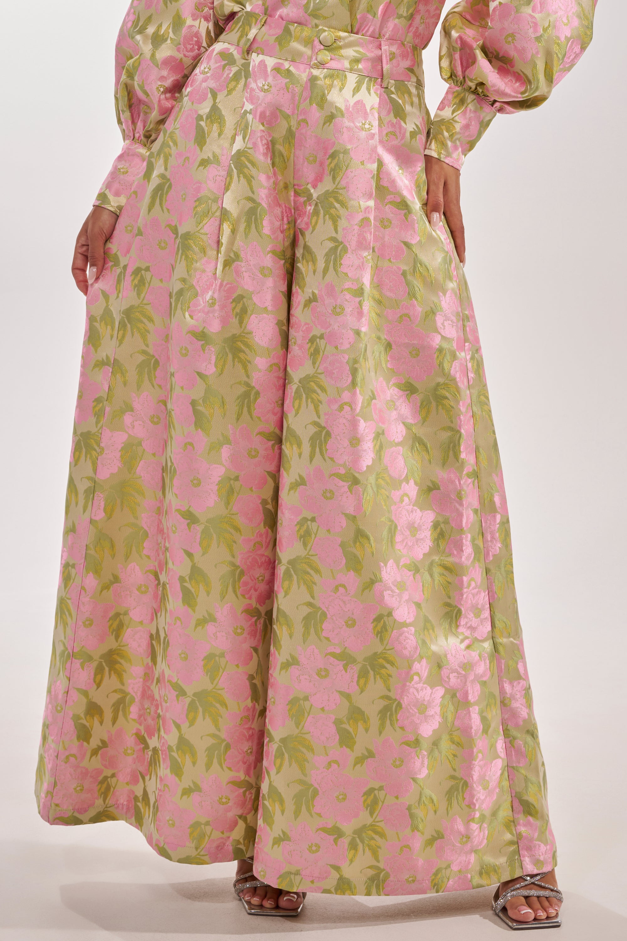 A person wears the OVER IT FLORAL TROUSER in pink and green floral print, paired with metallic silver open-toe heels. Only the lower half of their outfit is visible. 2