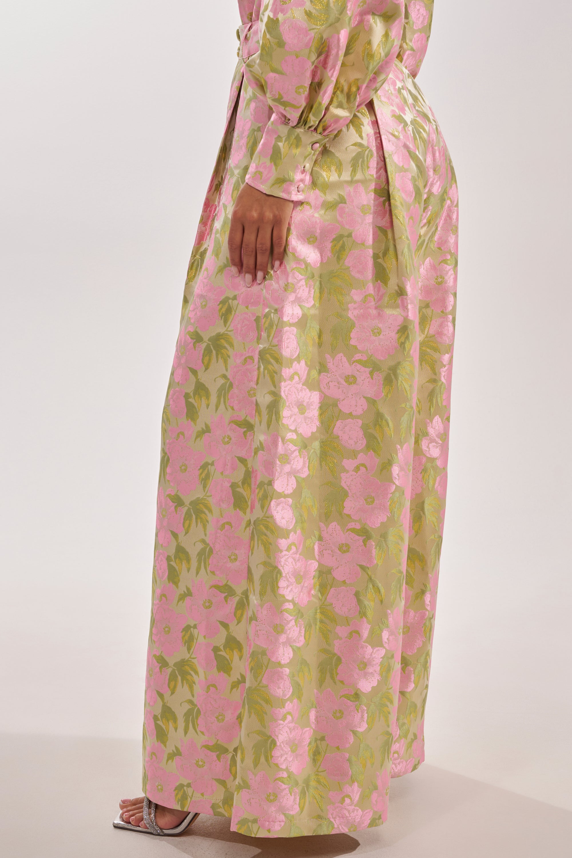 A person wears the OVER IT FLORAL TROUSER, featuring a high-rise waist and a light green and pink floral pattern, paired with strappy heeled sandals. Only the lower half of the body is shown. 3