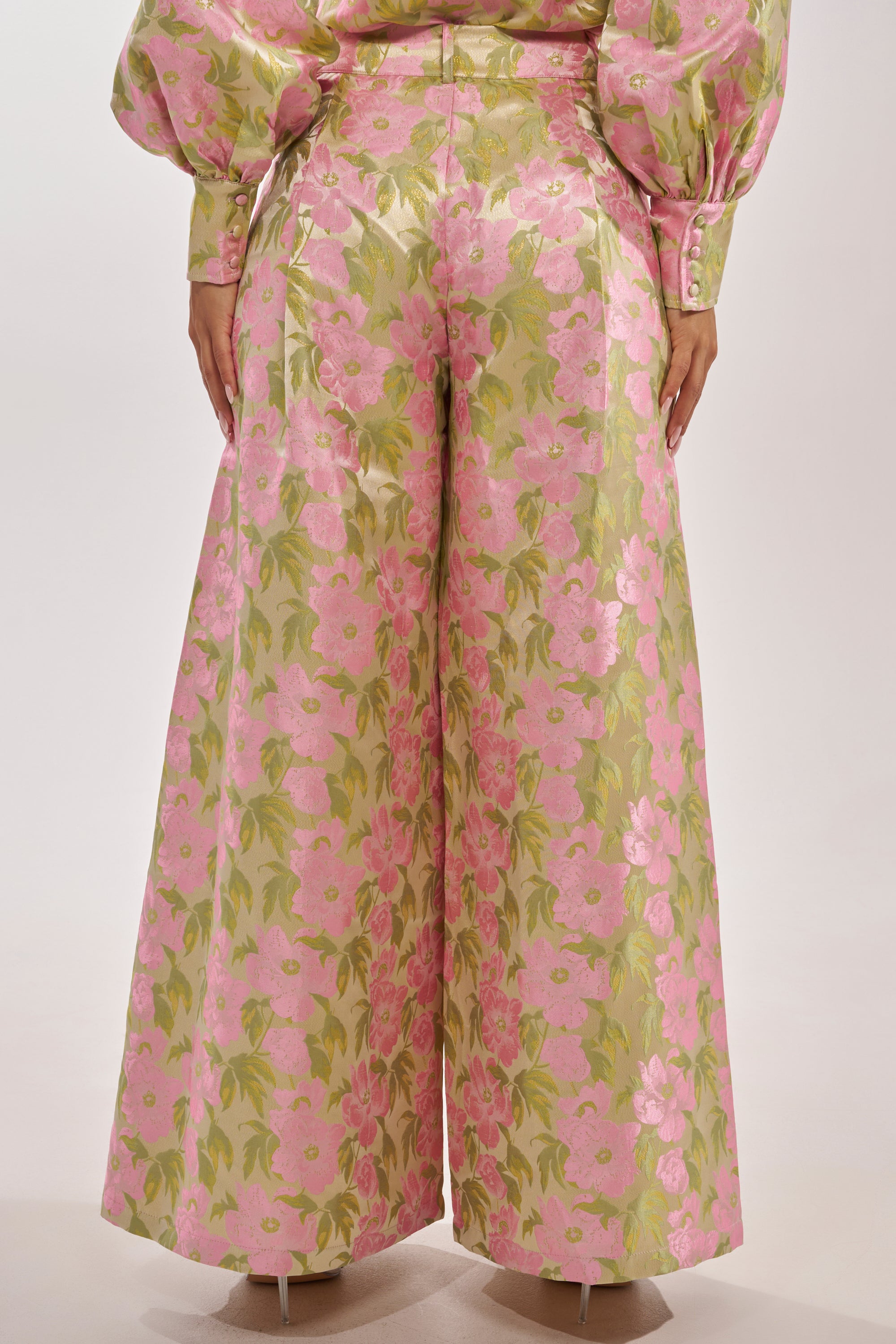 Someone is seen from behind wearing the OVER IT FLORAL TROUSER—a high-rise, full-length wide leg pant with a shiny pink and green floral pattern—paired with a matching long-sleeve top. 4