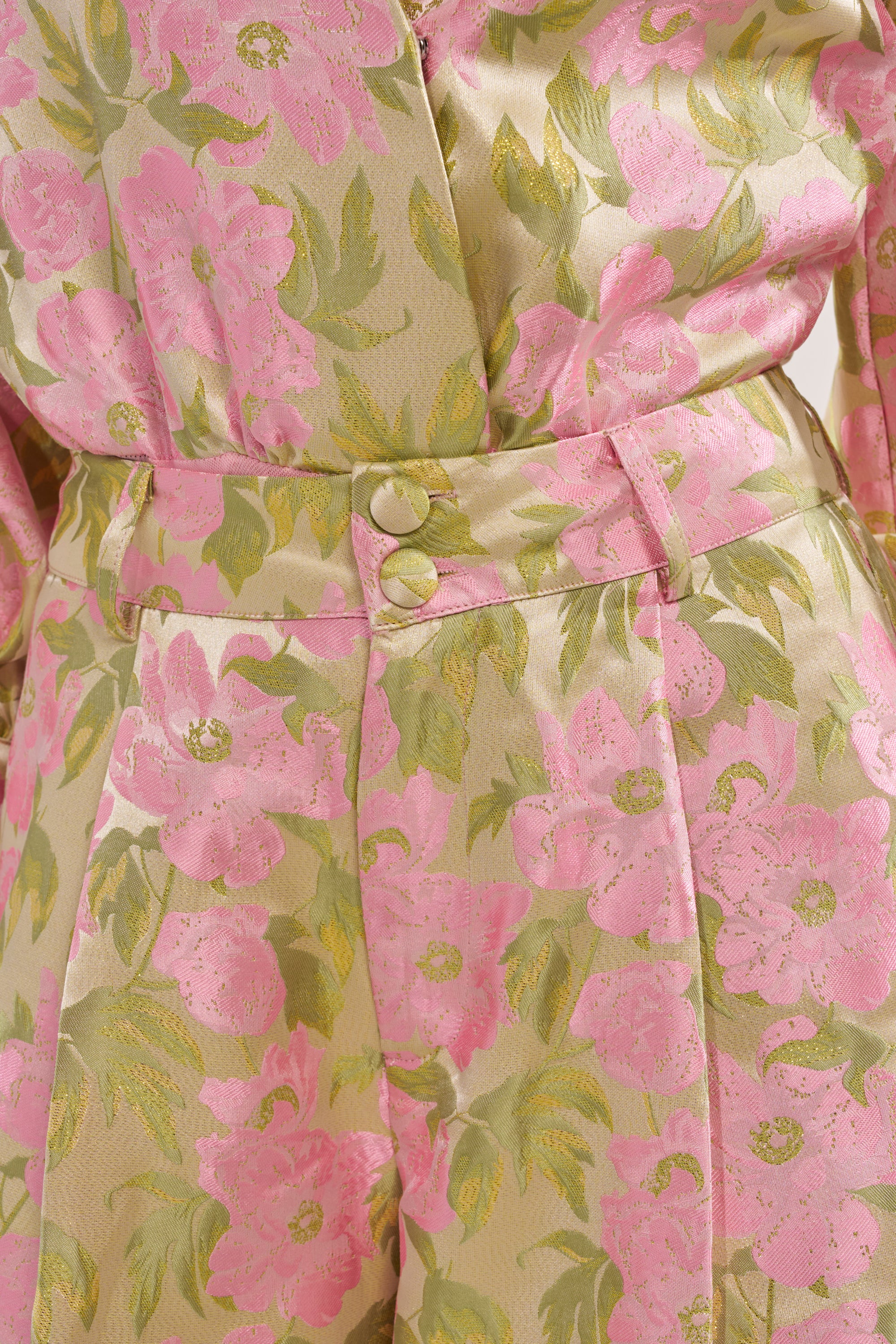Close-up of OVER IT FLORAL TROUSER shows shiny pink flowers and green leaves on beige fabric, highlighting a high-rise waist with two gold buttons and part of the matching top. 7