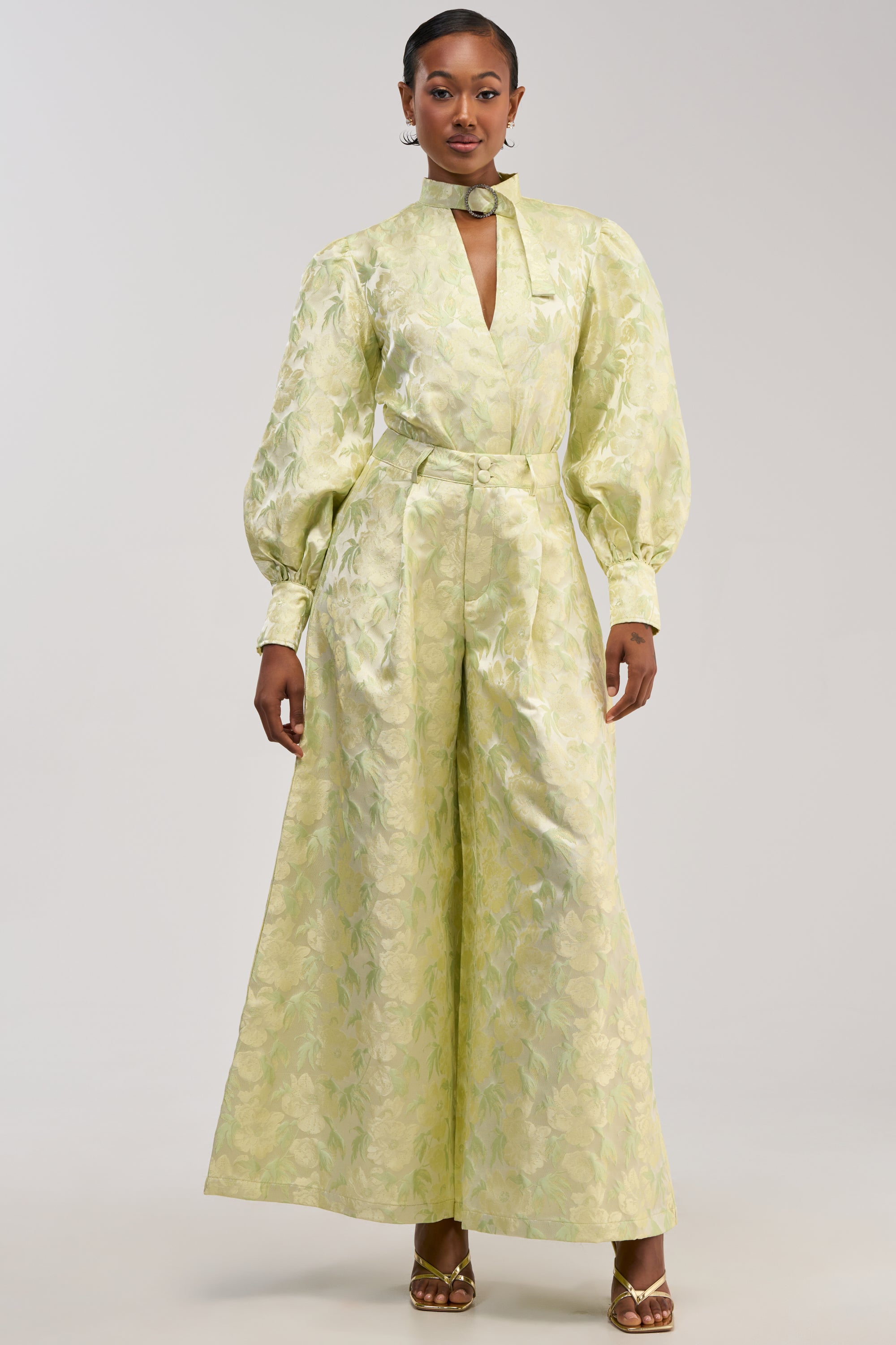 A woman stands against a plain background, wearing the LOOK THIS WAY SHINY PANT IN GREEN—a pair of light green, wide-leg pants that showcase an elegant jacquard-inspired pattern—styled with a matching long-sleeve top featuring puff sleeves, a high neck, and a coordinating belt for a polished look.