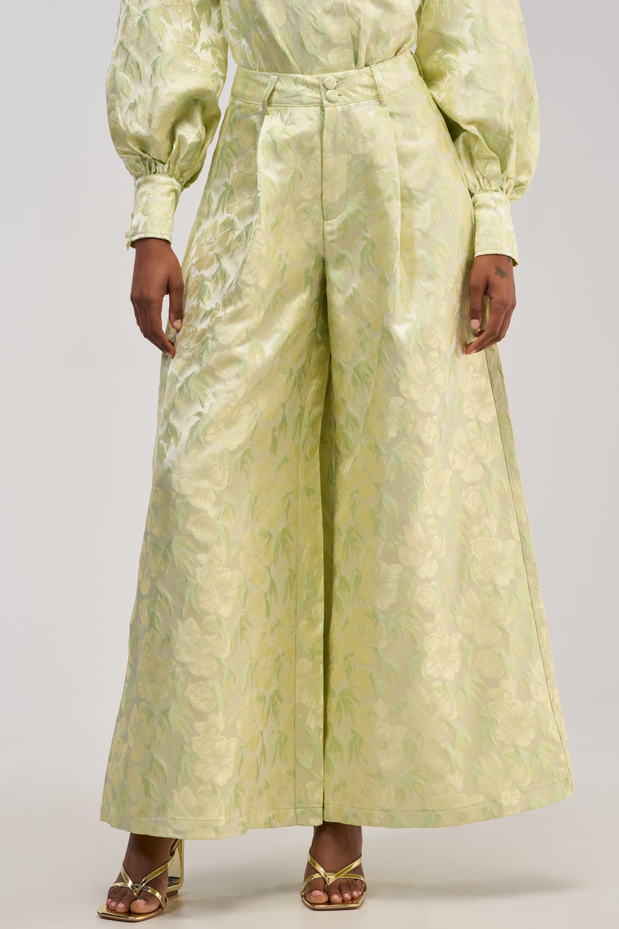 A person wearing the LOOK THIS WAY SHINY PANT IN GREEN—high-waisted, wide-leg jacquard holiday party pants in a light green floral pattern—paired with a matching long-sleeve blouse and gold open-toe heeled sandals. The background is plain and light-colored.
