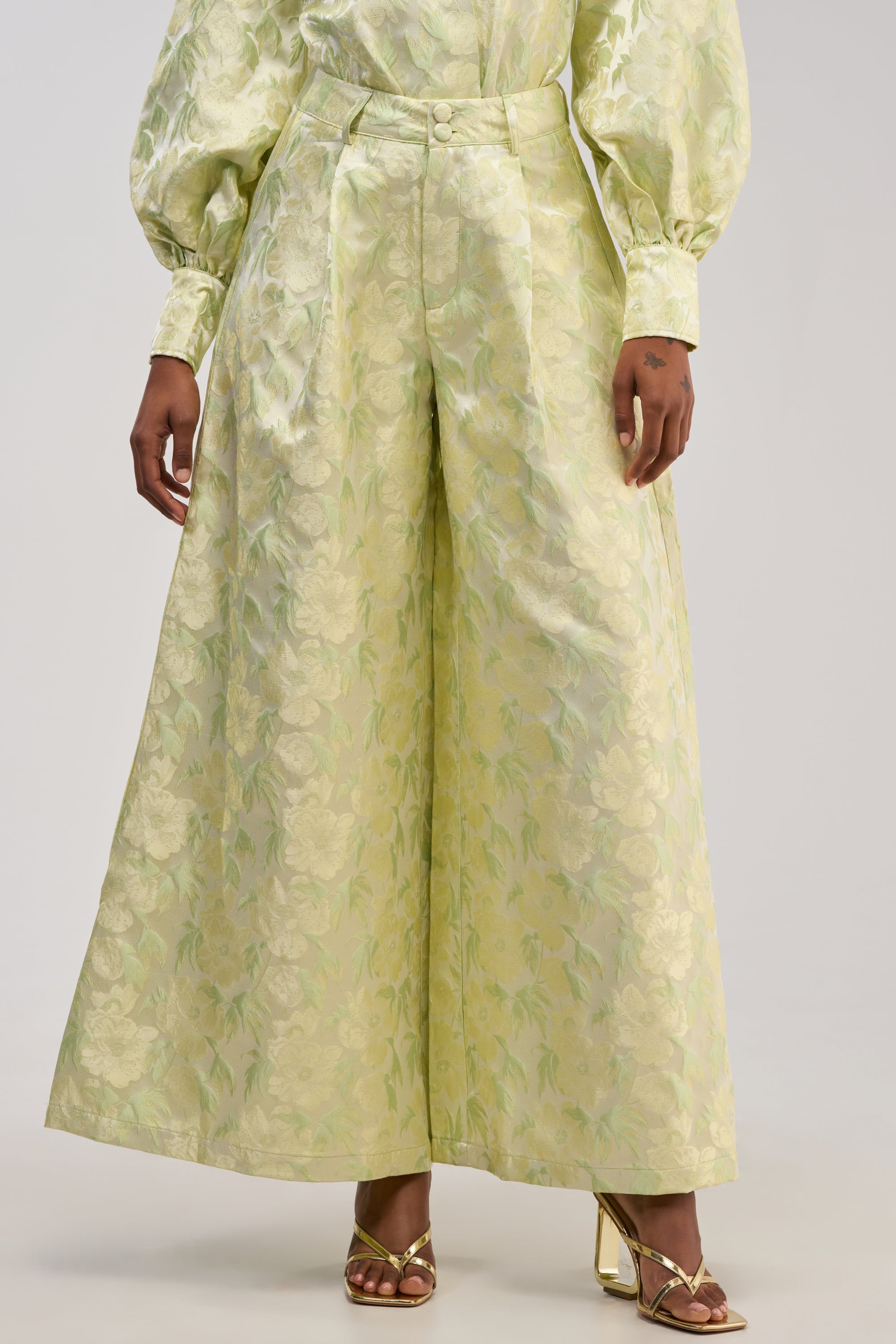 Wearing the LOOK THIS WAY SHINY PANT IN GREEN, styled with a matching puffy-sleeve shirt in pale yellow-green floral jacquard and gold open-toe heels.