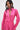 A woman models the LOOK THIS WAY SHINY BODYSUIT in hot pink metallic fabric with long sleeves, a high collar, and deep V-neck, paired with large dangling earrings, against a plain light background.