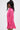 A woman in profile wears the LOOK THIS WAY SHINY BODYSUIT by AKIRA Label—a bright pink, long-sleeve collared piece. She has long, wavy dark hair and statement earrings. The background is plain white.