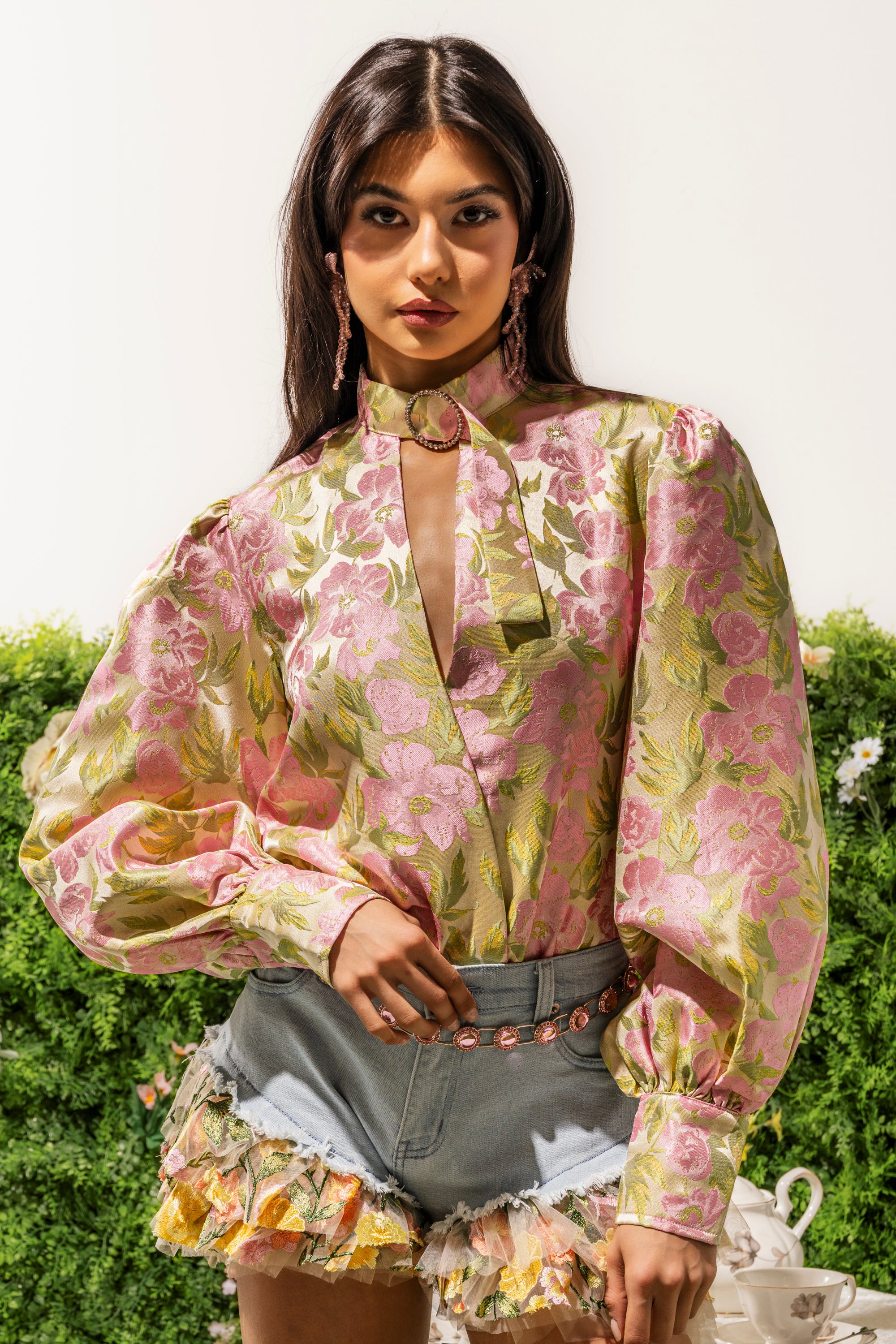 Wearing the OVER IT FLORAL BODYSUIT with puff long sleeves and a chic high neck, she pairs it with ruffled denim shorts and poses confidently by greenery, showcasing the floral print and keyhole detail. 0