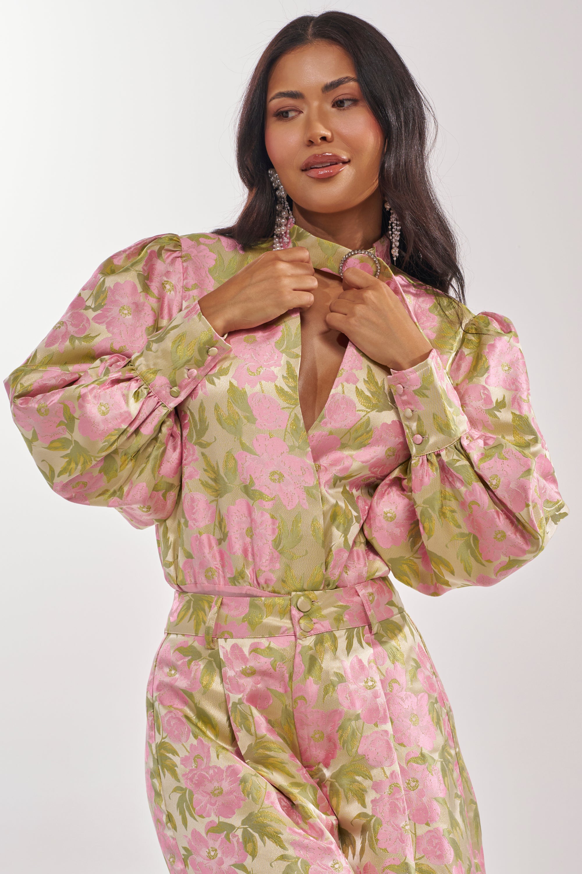 A woman with long dark hair and statement earrings confidently adjusts the collar of the OVER IT FLORAL BODYSUIT, a shiny pink and green floral print style, while posing against a light background. 2