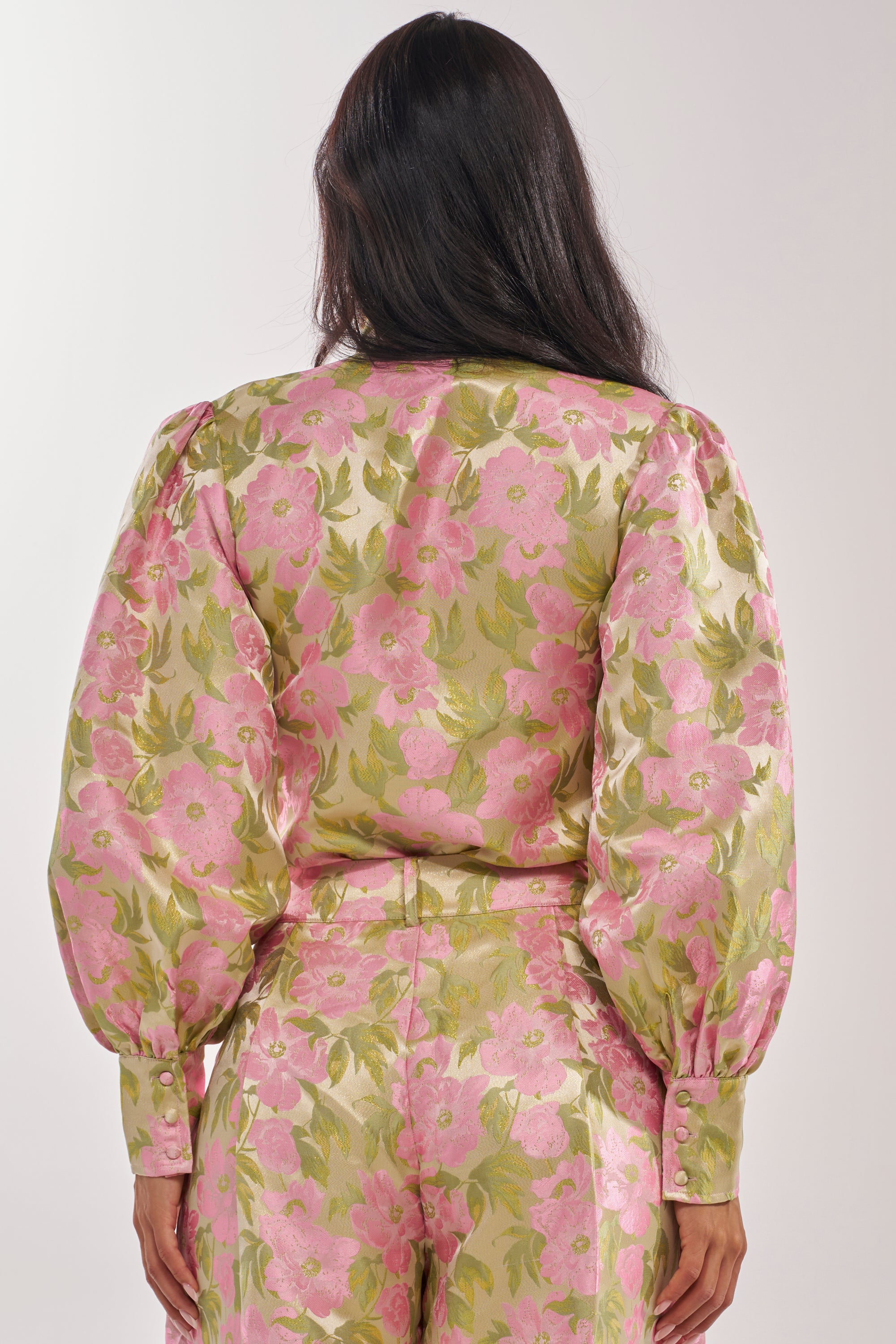 A person with long dark hair is seen from behind, wearing the OVER IT FLORAL BODYSUIT, which features shiny pink and green floral print, metallic details on puffed long sleeves, and high-waisted pants. 5