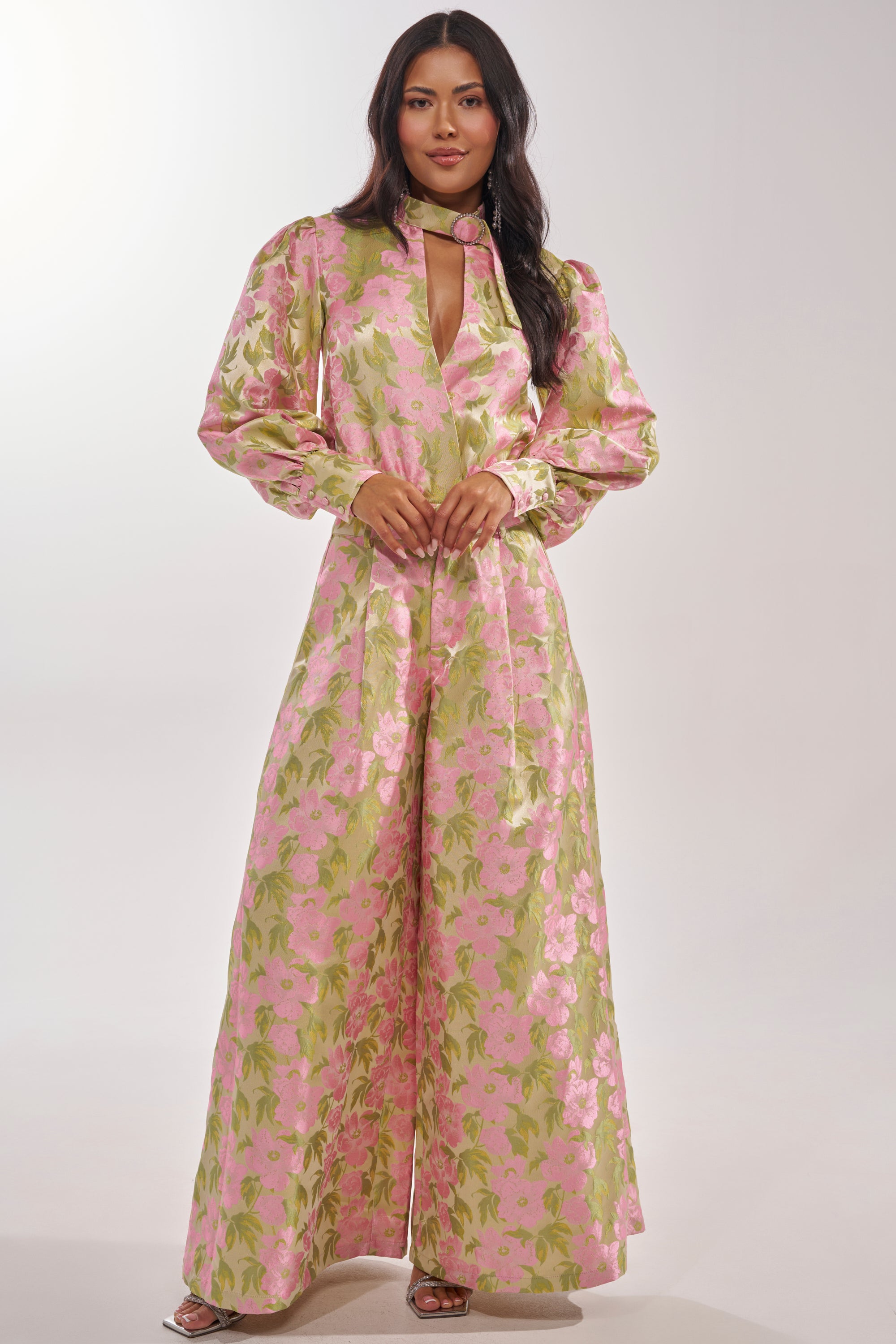 A woman with long dark hair models the OVER IT FLORAL BODYSUIT—a long-sleeve jumpsuit with a pink and green floral pattern, metallic details, high neckline, and chest keyhole—posing against a plain background. 6