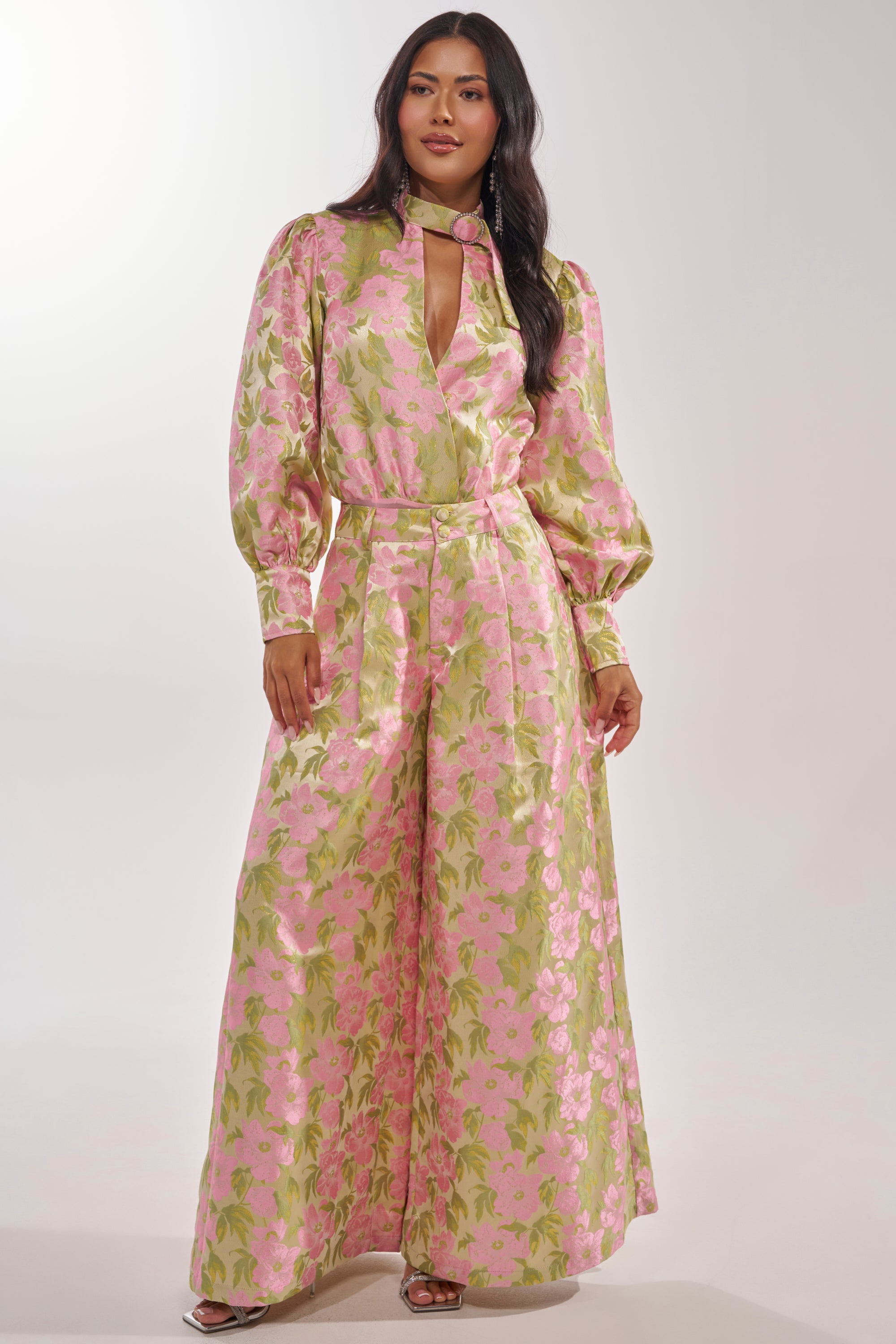 A woman models the OVER IT FLORAL BODYSUIT—shiny, long-sleeve, with a keyhole neckline, pink and green floral print, rhinestone buckle, and wide-leg pants—against a plain white background. 8