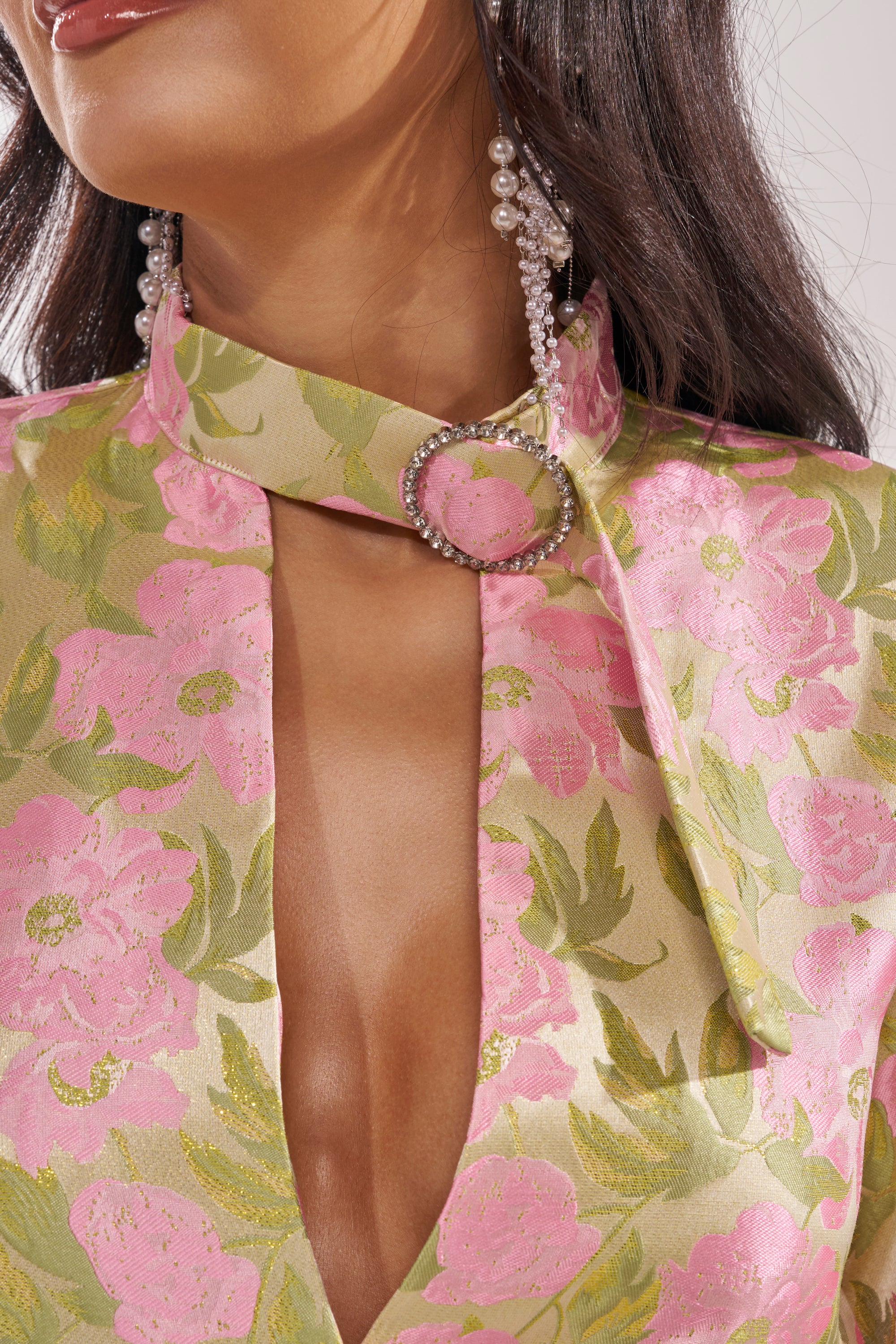 A woman models the OVER IT FLORAL BODYSUIT, featuring a pink and green floral print, deep neckline, and collared design with metallic fasteners. She pairs it with long dark hair and dangling pearl earrings. 9