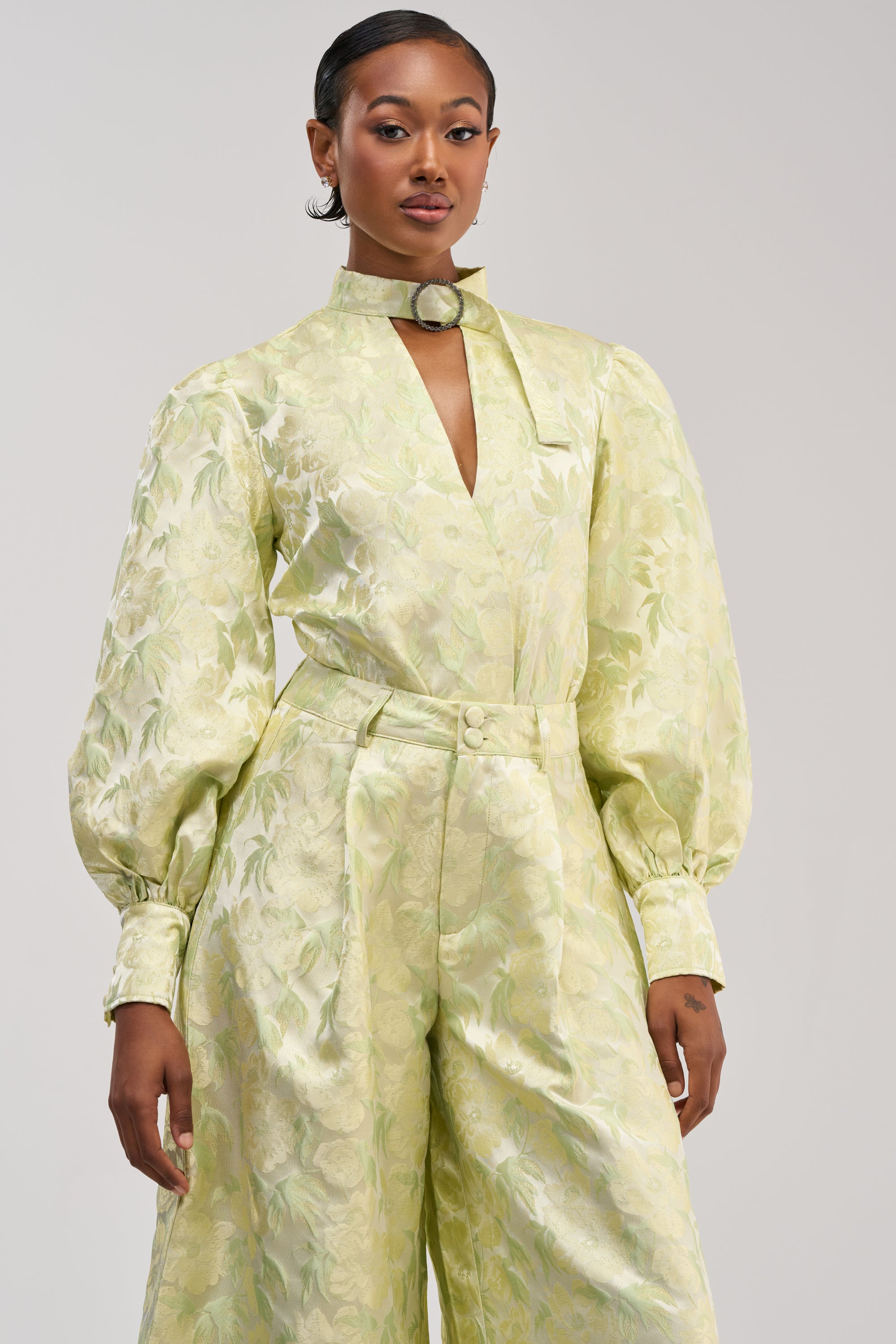 A woman wearing the LOOK THIS WAY Shiny Bodysuit in green, featuring jacquard fabric, puffed sleeves, and a choker collar, stands confidently against a plain background.