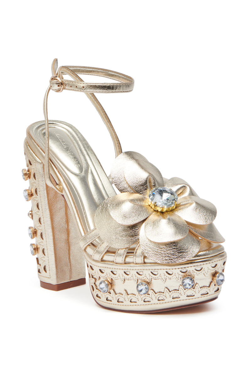 The AZALEA WANG PIXLEY GOLD EMBELLISHED PLATFORM SANDAL features metallic faux leather, an ankle strap, chunky heel, a large metallic flower with central gemstone, and smaller round gems on the platform and heel.
