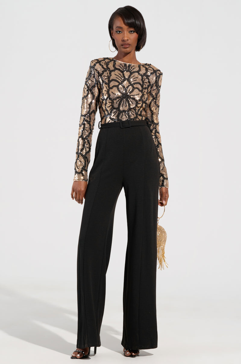 A woman stands confidently in a party-ready ensemble featuring the BETTER OFF SOLO JUMPSUIT against a neutral background. This outfit flaunts long sleeves with a gold and black sequin floral pattern. She complements her look with a small gold purse and stylish heels, exuding elegance in every detail.