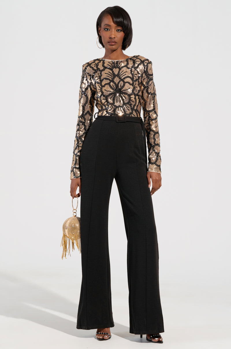 A person stands confidently, ready to party in the BETTER OFF SOLO JUMPSUIT featuring a gold sequined top with an intricate floral pattern and high-waisted black wide-leg pants. They complement their look with a round decorative handbag adorned with fringes and sleek black open-toe heels.