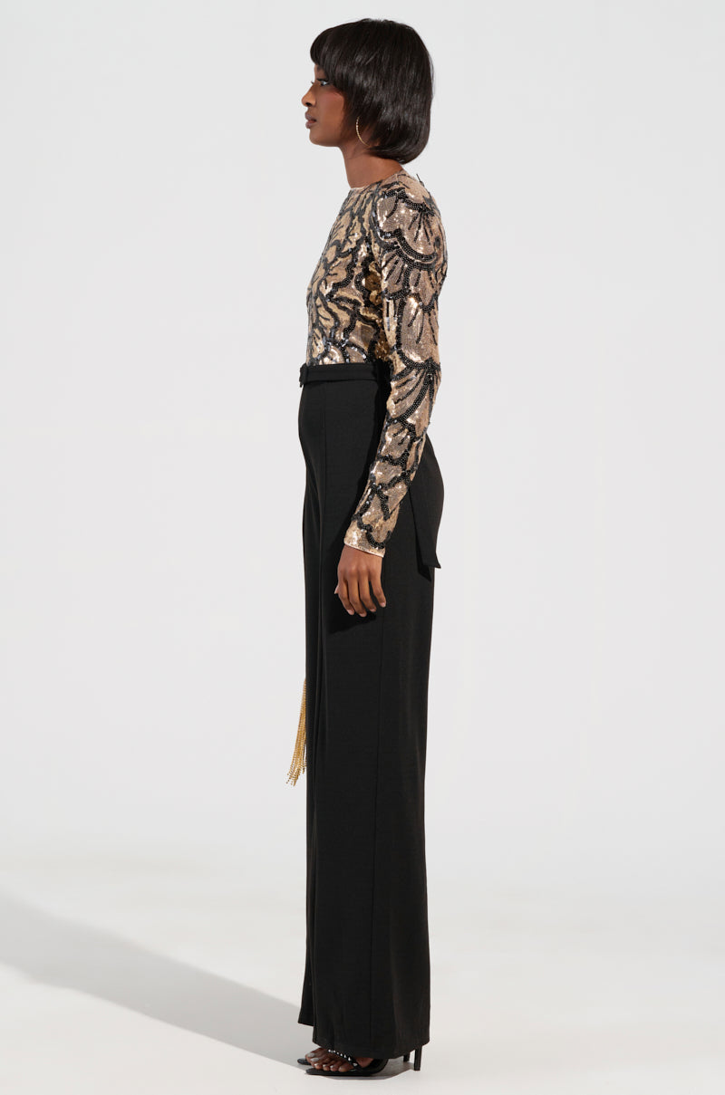 The image showcases an individual in profile, dressed in a "BETTER OFF SOLO" jumpsuit featuring intricate metallic patterns that evoke sequin floral designs. Their high-waisted black trousers and heels enhance the party-ready vibe, while their short, dark hair stands out against the plain, neutral background.
