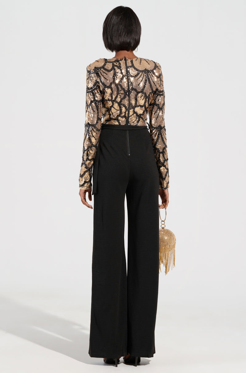 A person is seen from behind, wearing the BETTER OFF SOLO JUMPSUIT, featuring a shimmering gold and black sequin floral pattern design. Party-ready, they hold a small gold tassel purse.