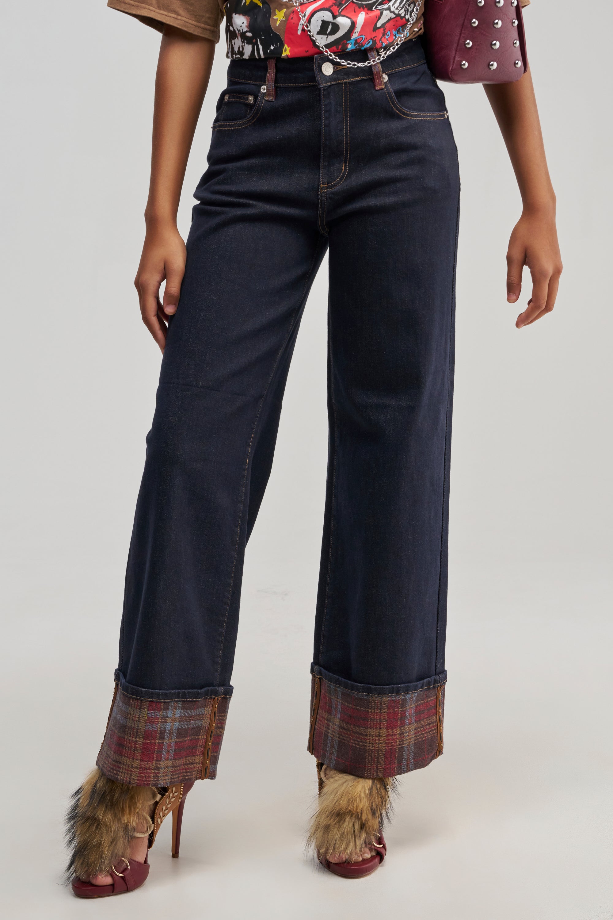 Only the lower half of the body is visible: a person wearing THE COOL PLAID JEANS STRAIGHT FIT, featuring dark wash denim with wide legs, plaid detailing, and brown fur cuffs. They are also wearing maroon high heels and a colorful top.