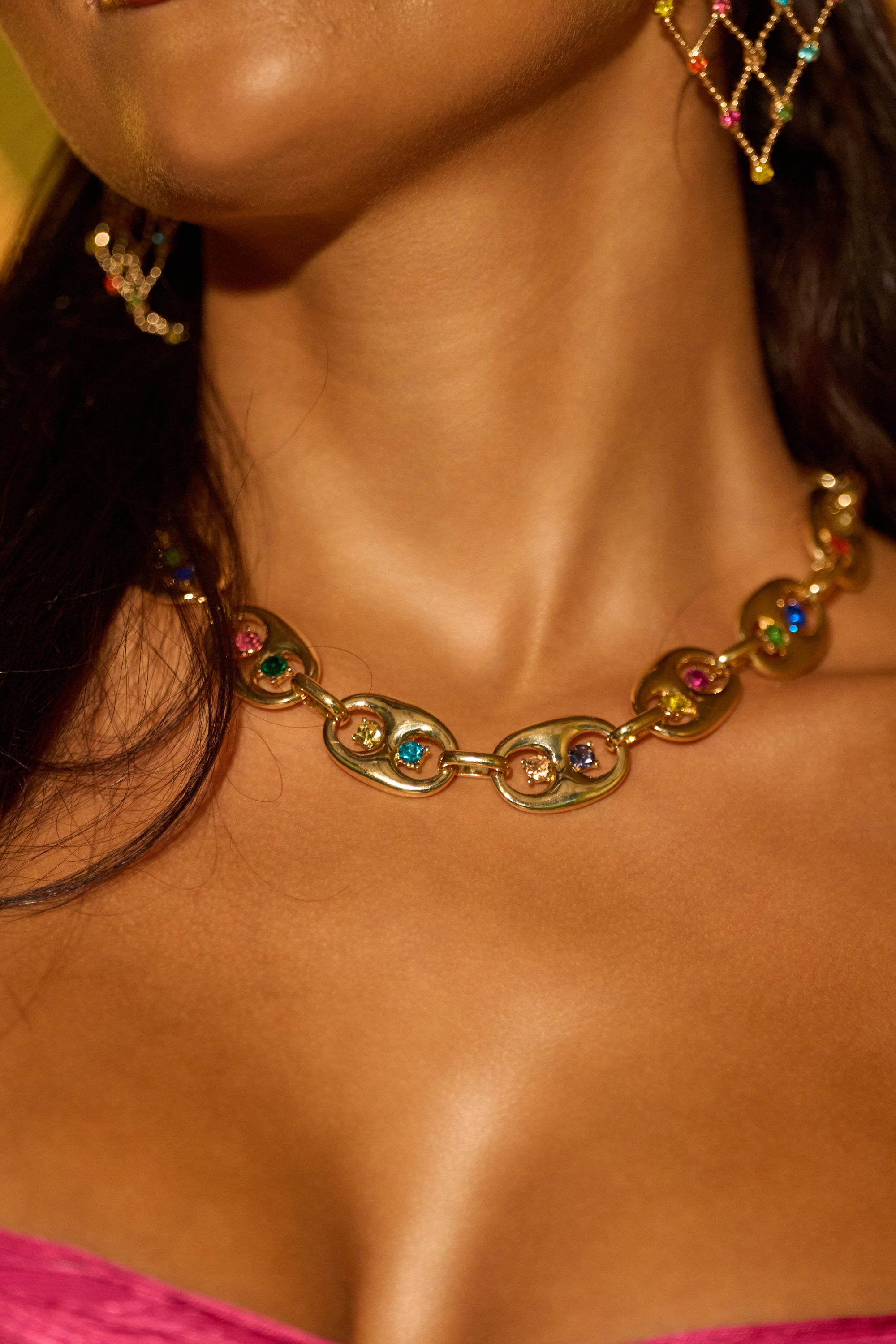 A close-up of a woman with smooth brown skin wearing the A GOOD TIME NECKLACE, a chunky gold rhinestone necklace with colorful gemstones, and a netted gold earring. She pairs this festival jewelry style with a pink top. 0